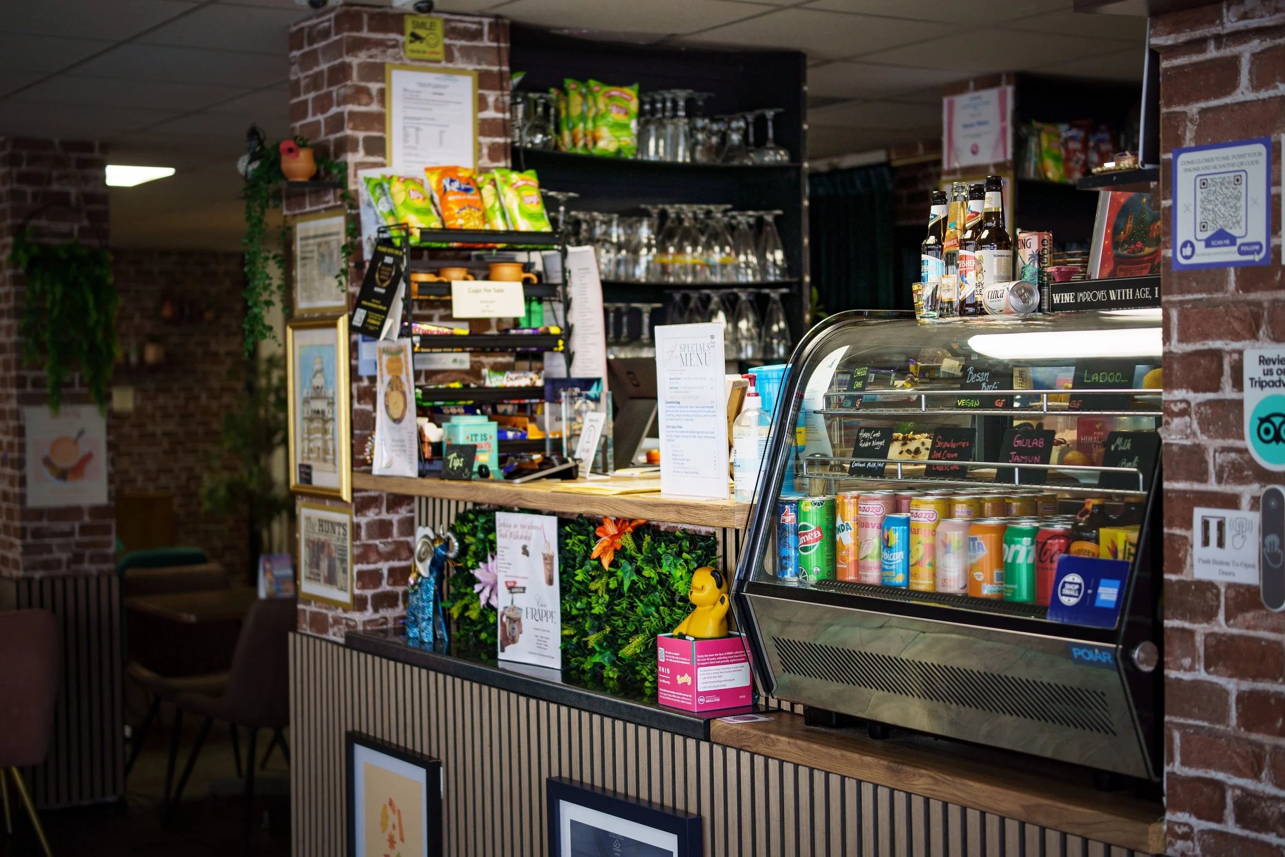 Interior of a cafe or convenience store with a brick counter, glass display case of canned and bottled drinks, shelves with glassware, snack bags, and various signs and decorations on the brick walls.