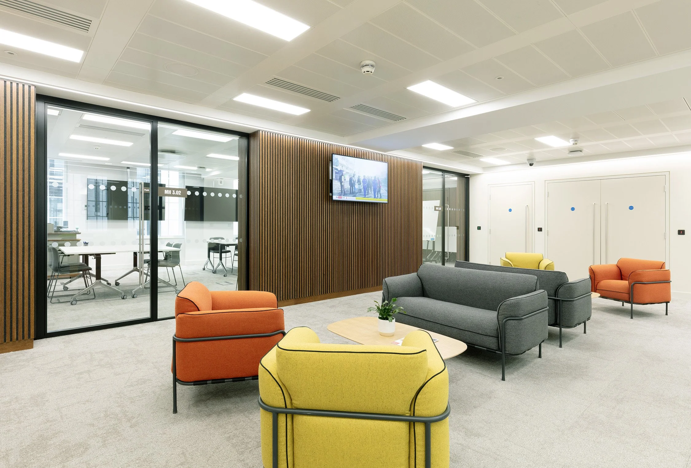 Modern office lounge area with colorful armchairs and couches in yellow, orange, and gray around a light wood coffee table with a potted plant, enclosed conference rooms with glass doors on the left, a wall-mounted TV on a wooden panel, and white wal