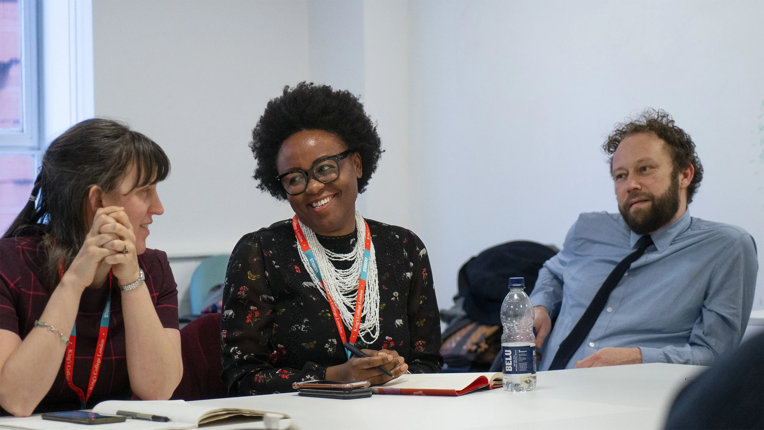 Three people sitting at a table engaged in a meeting, with notebooks, a water bottle, and a phone on the table.