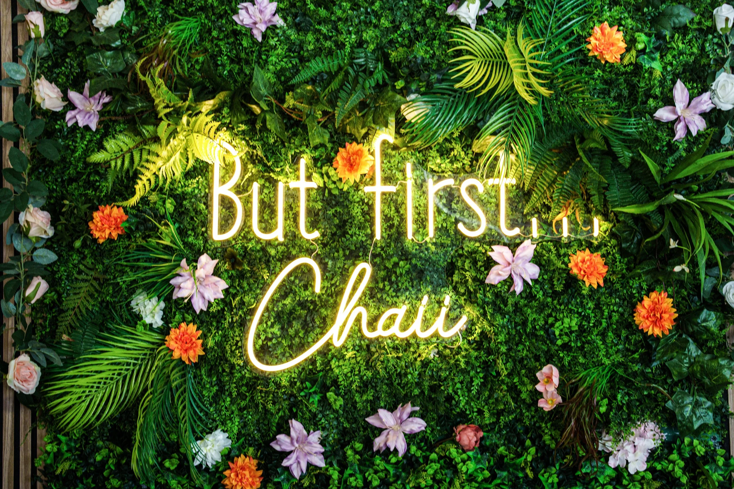 Neon sign that reads 'But first, chai' surrounded by green foliage and colorful flowers.