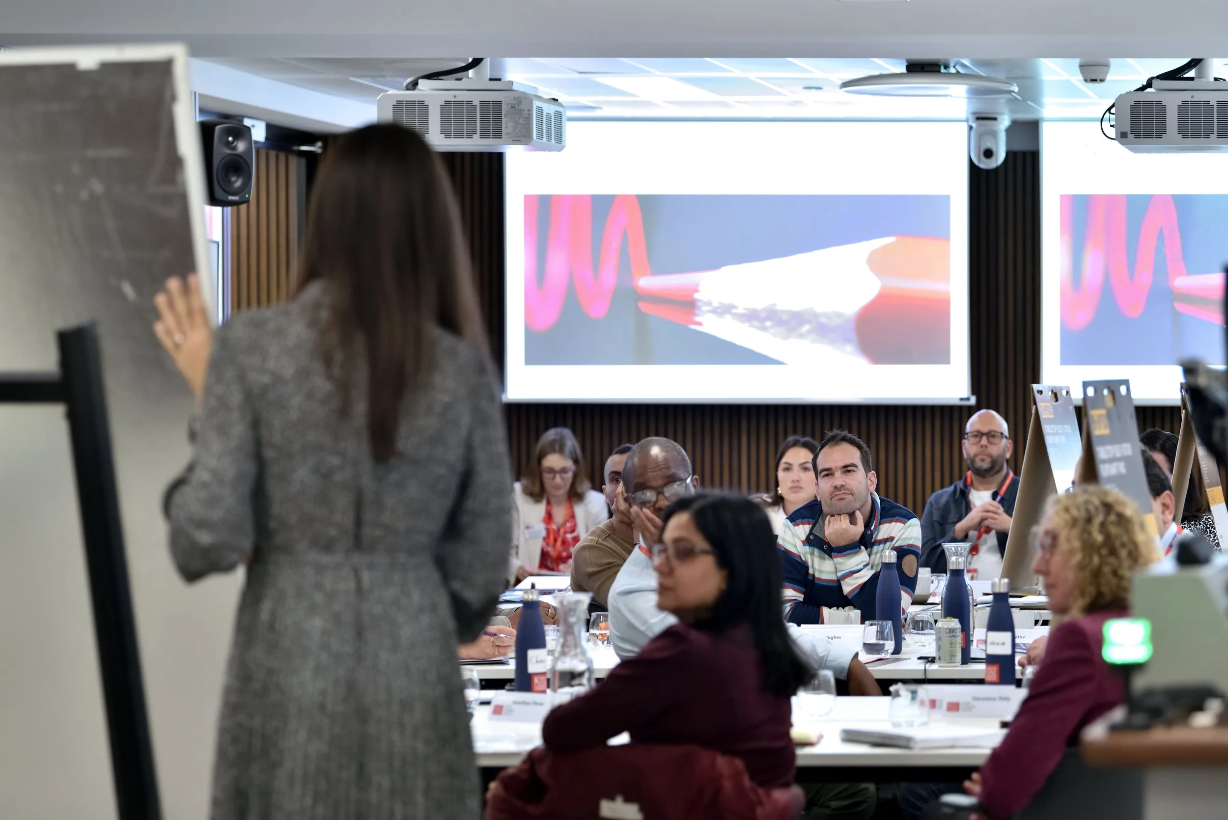 Woman giving presentation in front of a conference room with attentive attendees, large screen displaying abstract graphics.