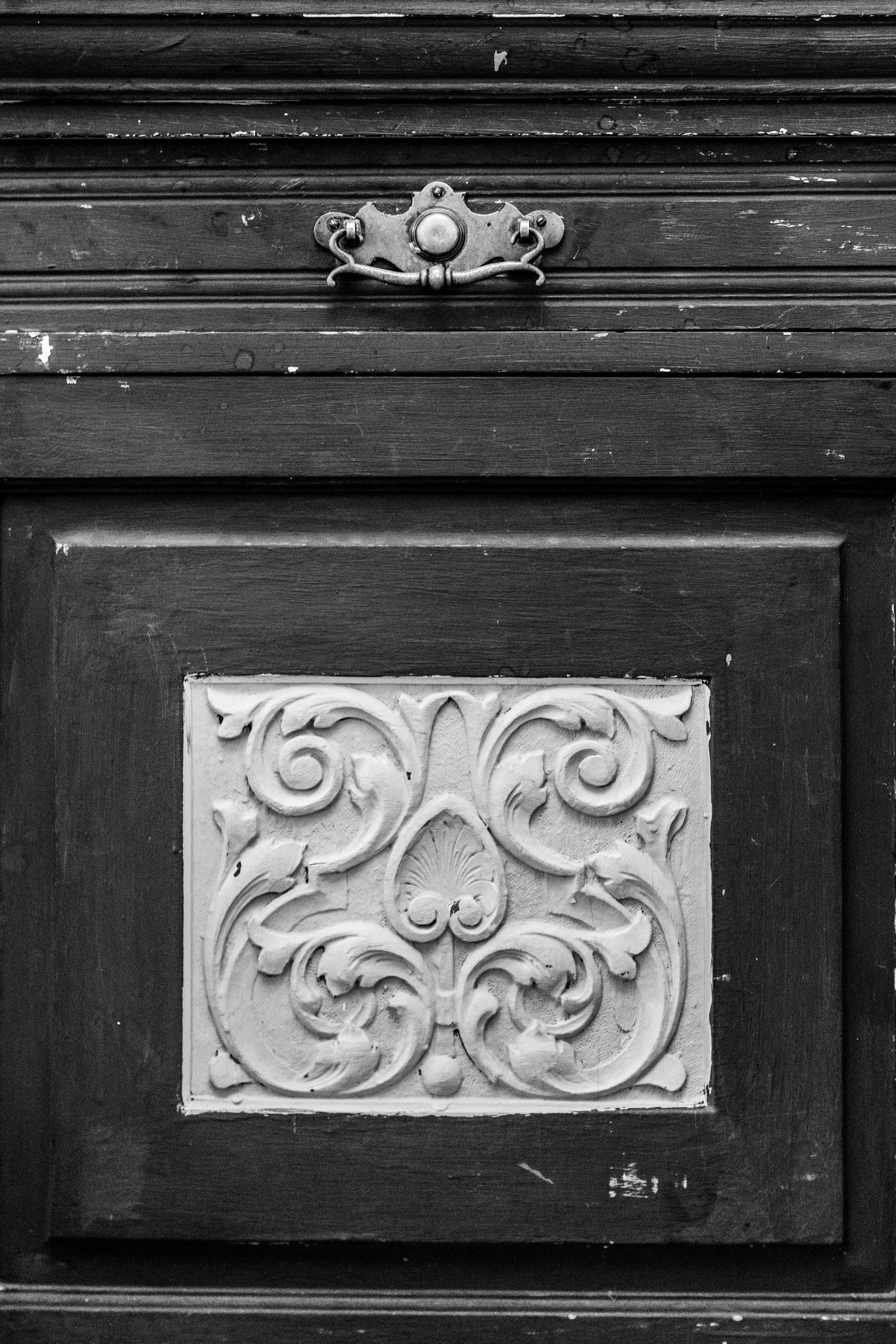 Black and white photo of a vintage wooden cabinet with an ornate decorative panel on the door and a metal latch above it.