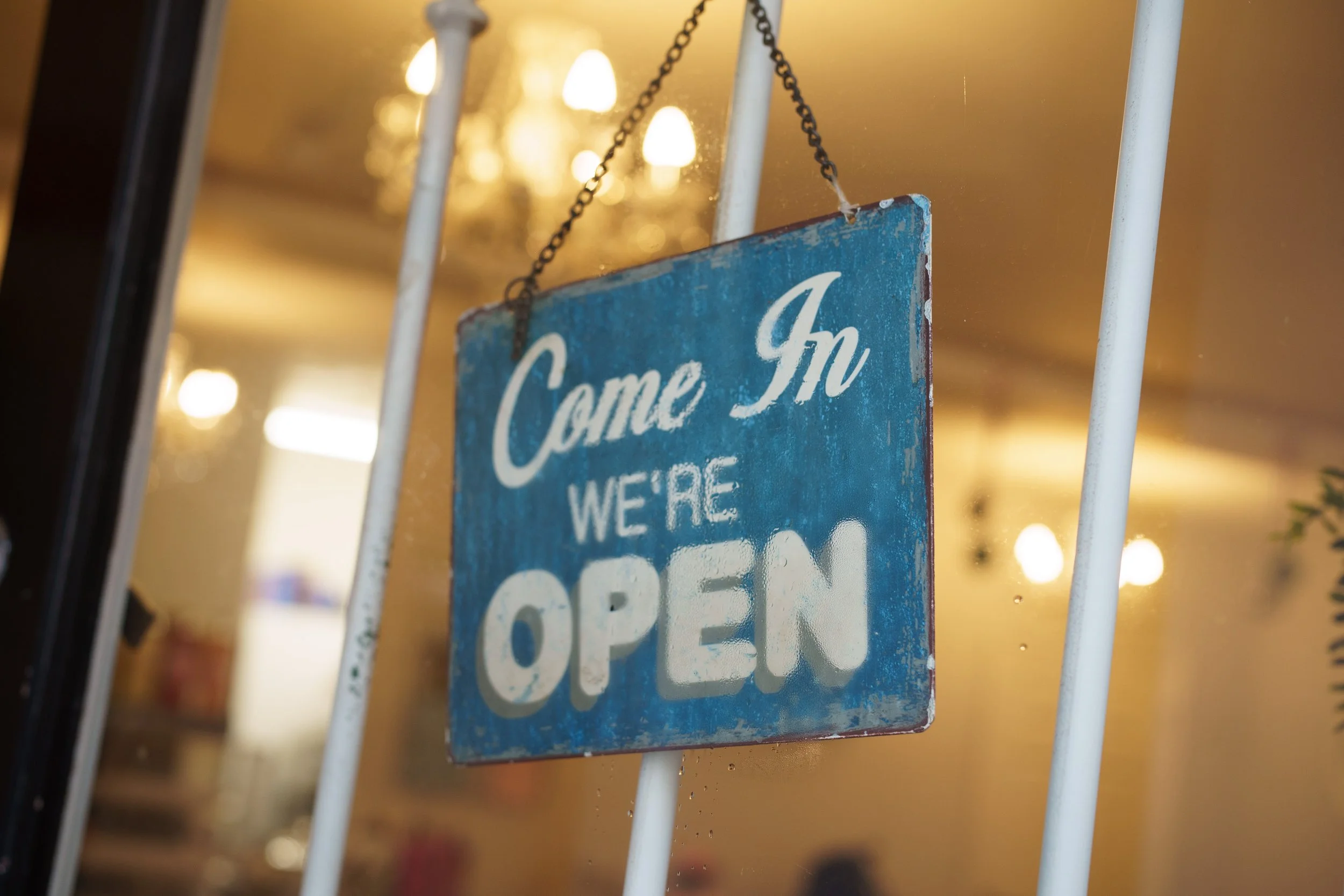 A blue, weathered sign hanging on a glass door that reads 'Come In We're OPEN' in white text.