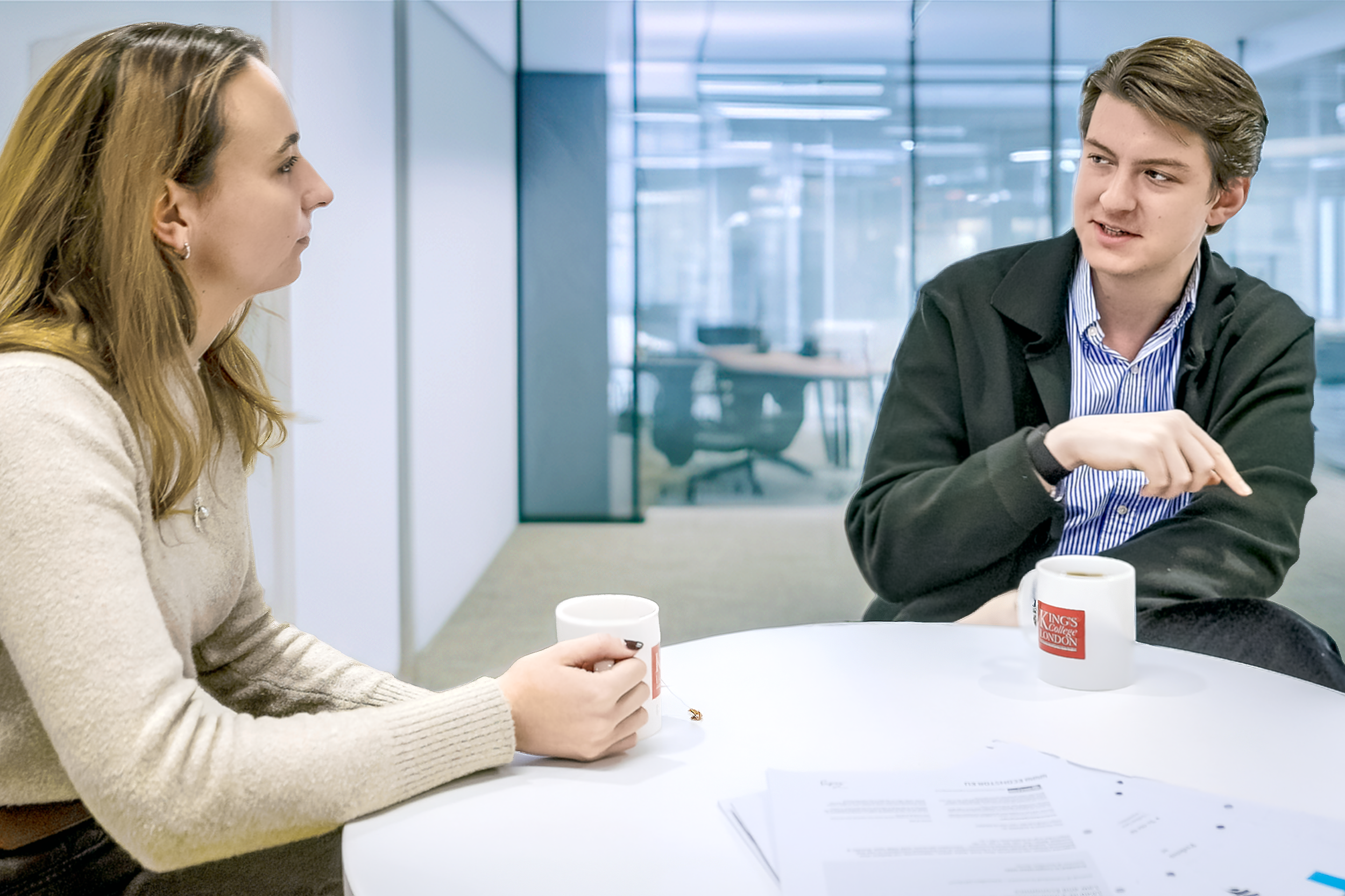 Two people having a conversation at a white table in an office setting, with coffee mugs and papers on the table.