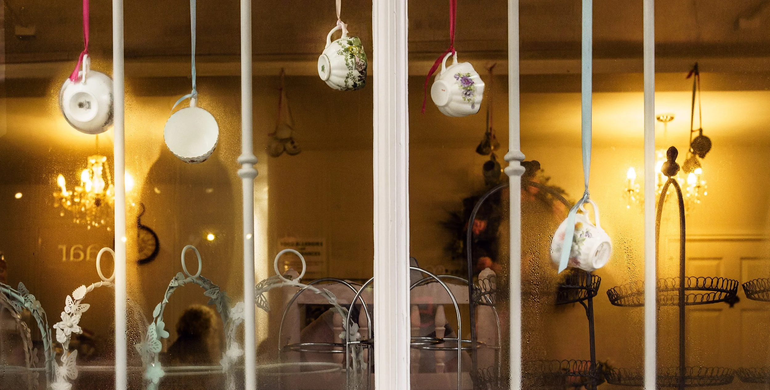 Decorative teacups hanging on white bars in front of a glass window, with a warm interior lighting and chandeliers inside.