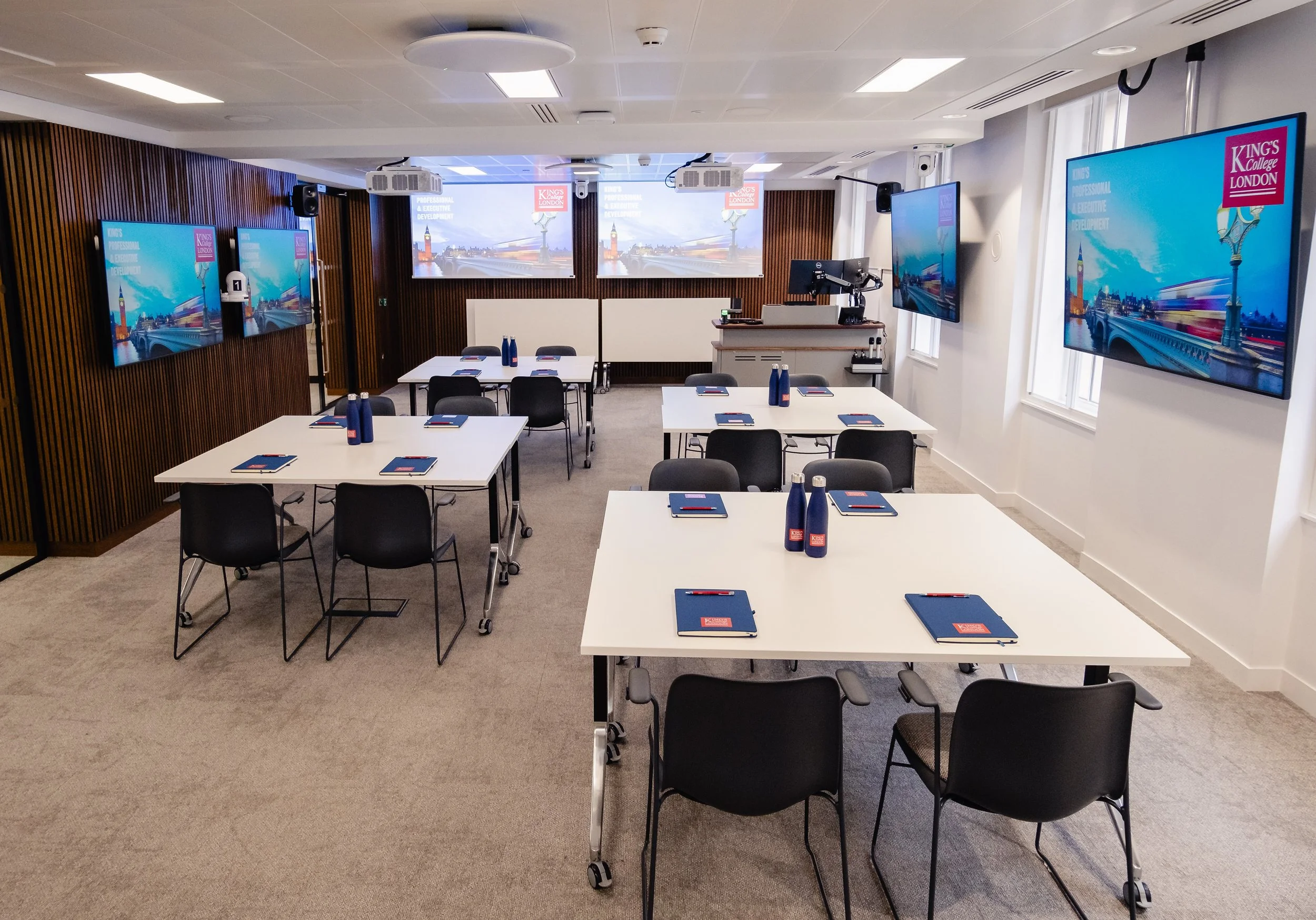 Modern conference room with white tables arranged in rows, equipped with notebooks, pens, and bottles of water. Multiple large screens display a presentation with the logo for King's College London. The room has wooden paneling on one wall, white wal