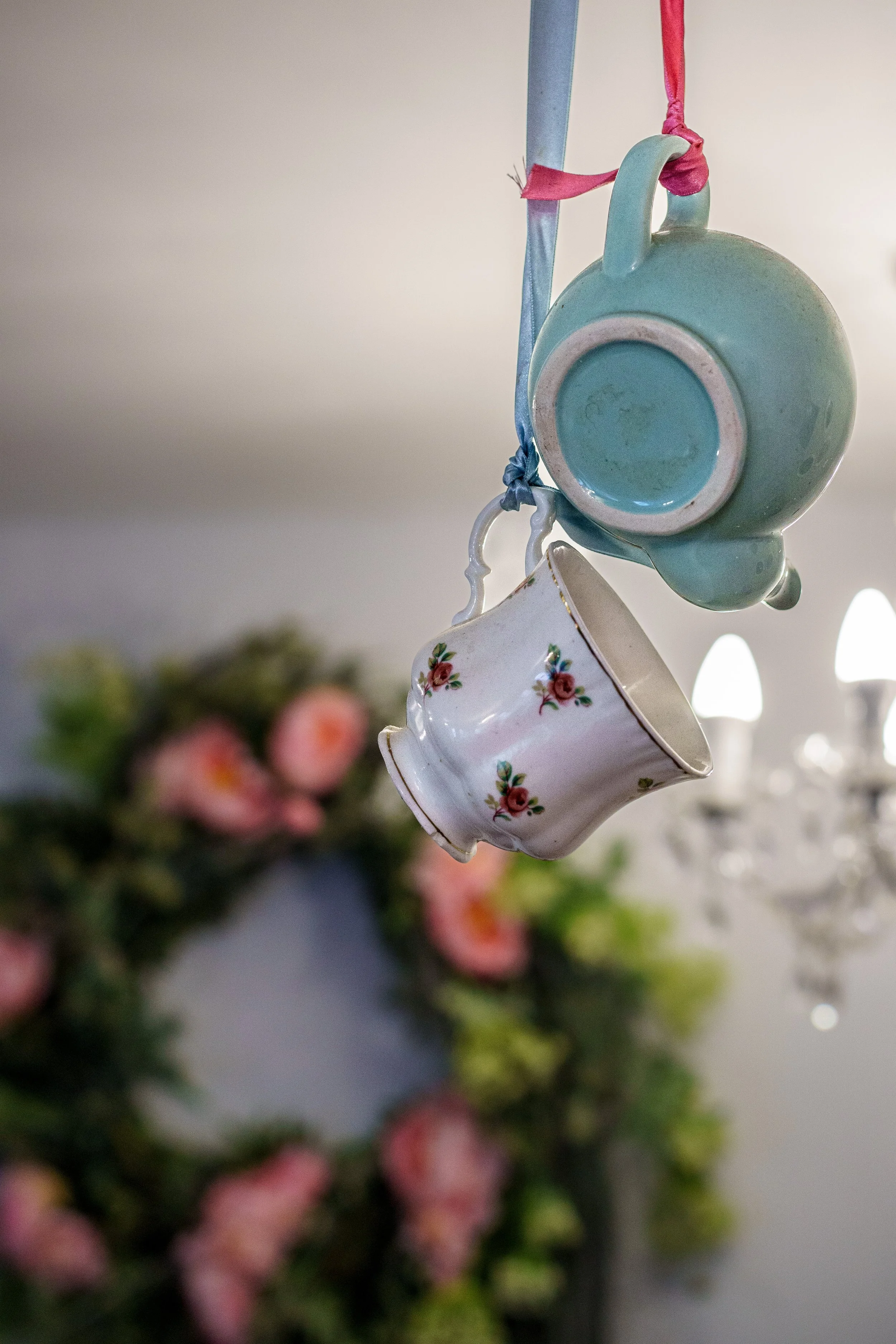 Hanging ceramic cups, one blue and one white with pink floral pattern, against a blurred background of pink flowers and a chandelier.
