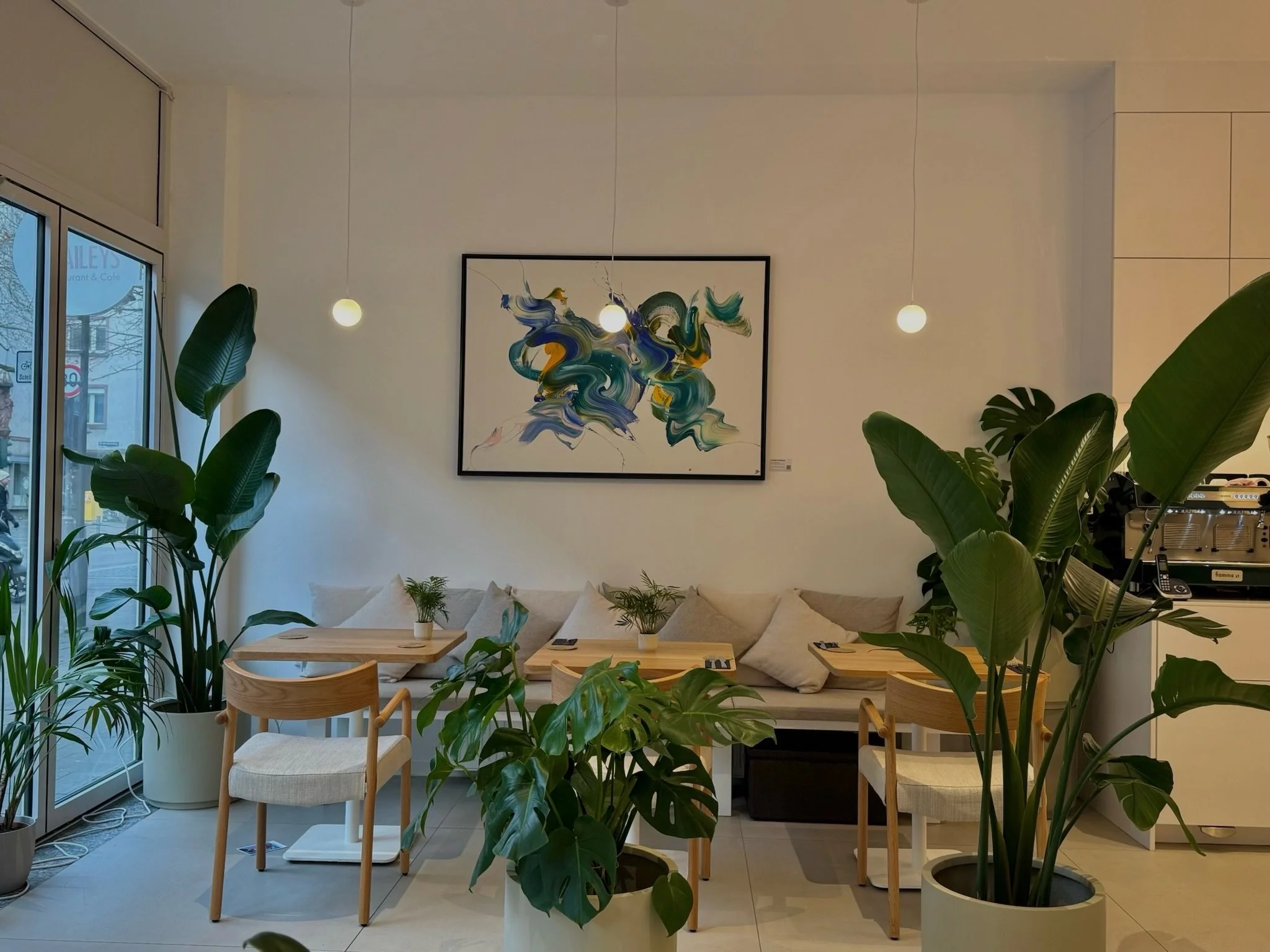 Gemütliches Café mit grünen Pflanzen, einem gemütlichen Sessel, einem Sofa mit Kissen, Holztischen, moderner abstrakter Kunst an der Wand, und warmem Licht durch Pendelleuchten.