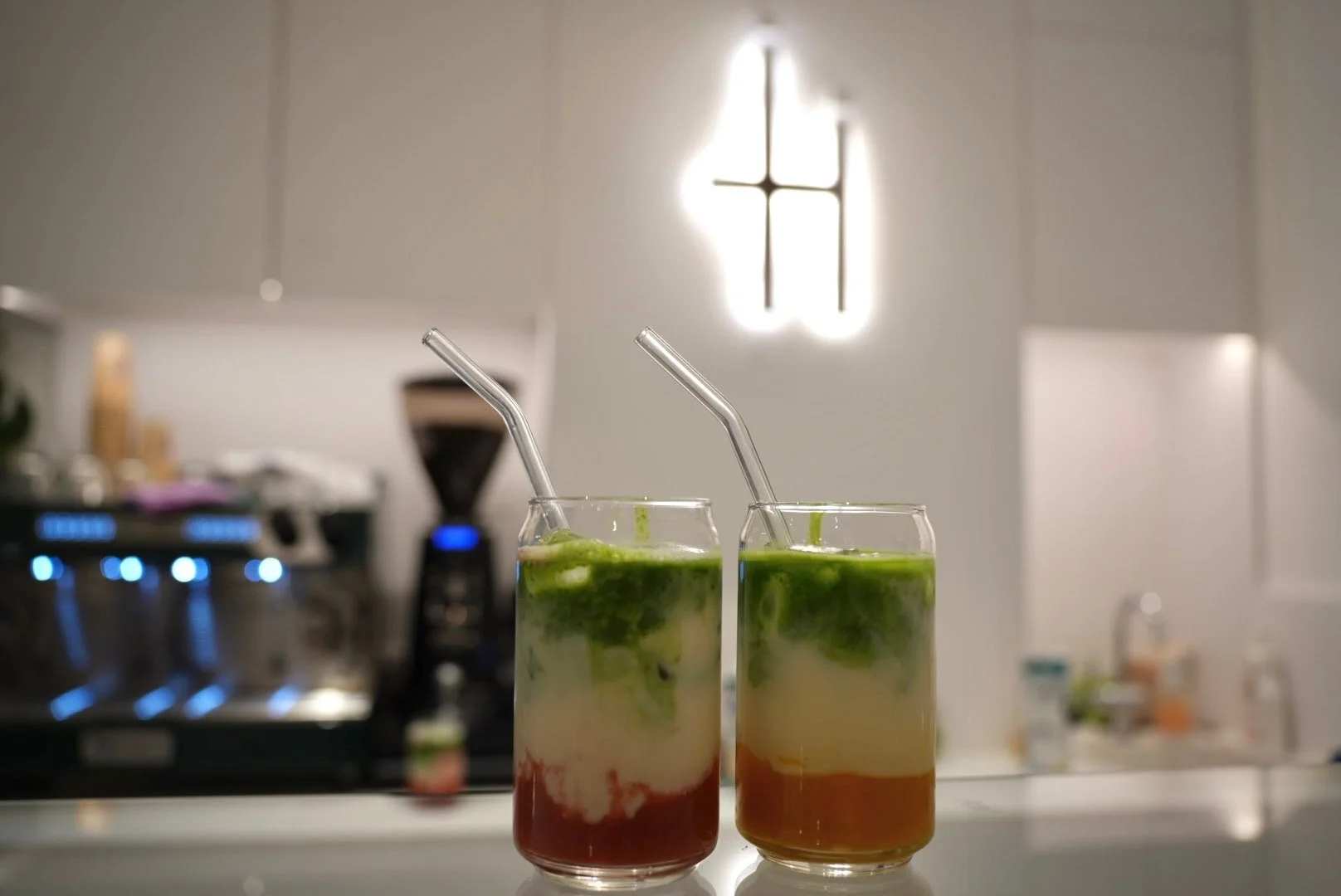 Zwei bunte Getränke mit Strohhalmen auf einer weißen Oberfläche, im Hintergrund eine Kaffeemaschine und eine moderne Leuchte mit Kirchenfenster-Design an der weißen Wand.