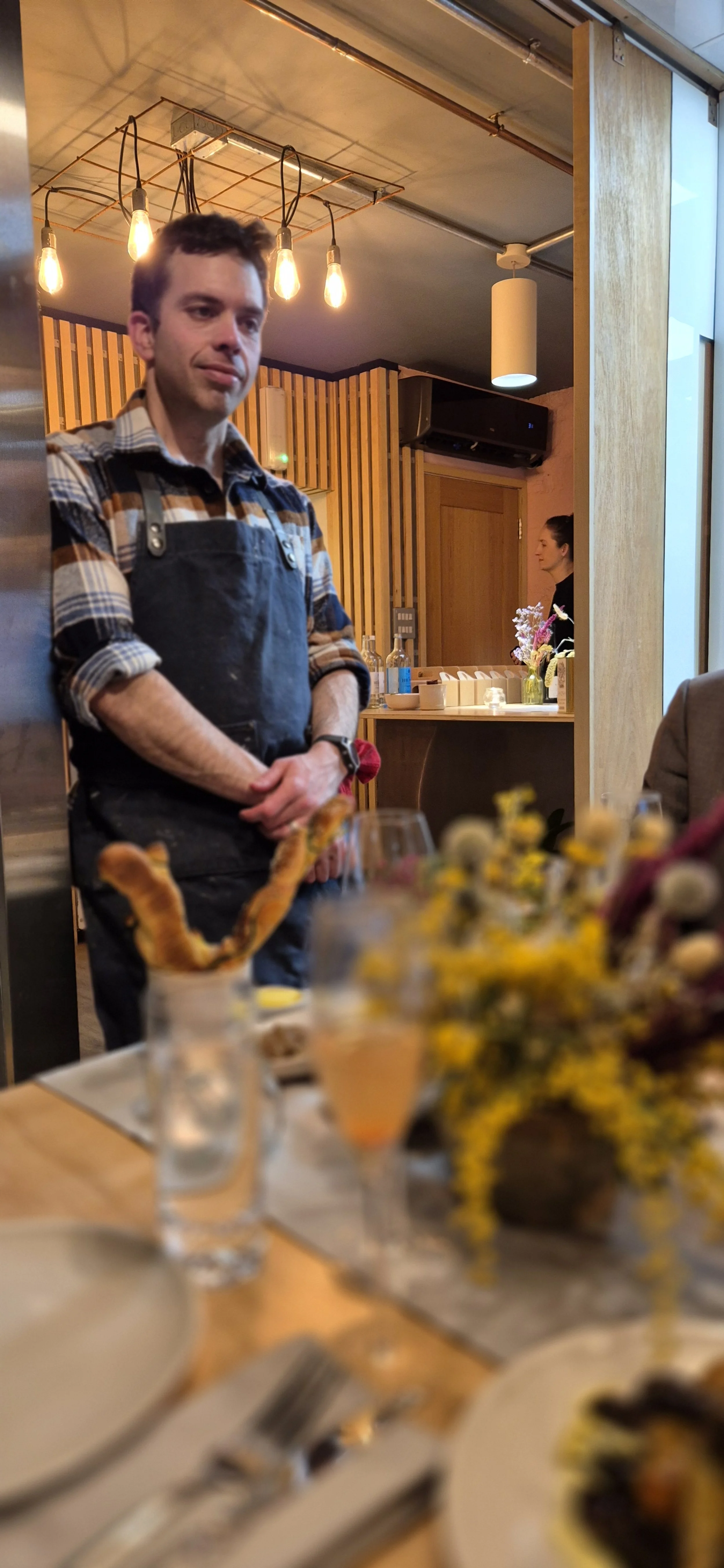 Restaurant interior with a male server in an apron standing beside a table set with plates, cutlery, and a vase of flowers. Warm lighting and wood paneling are visible in the background.