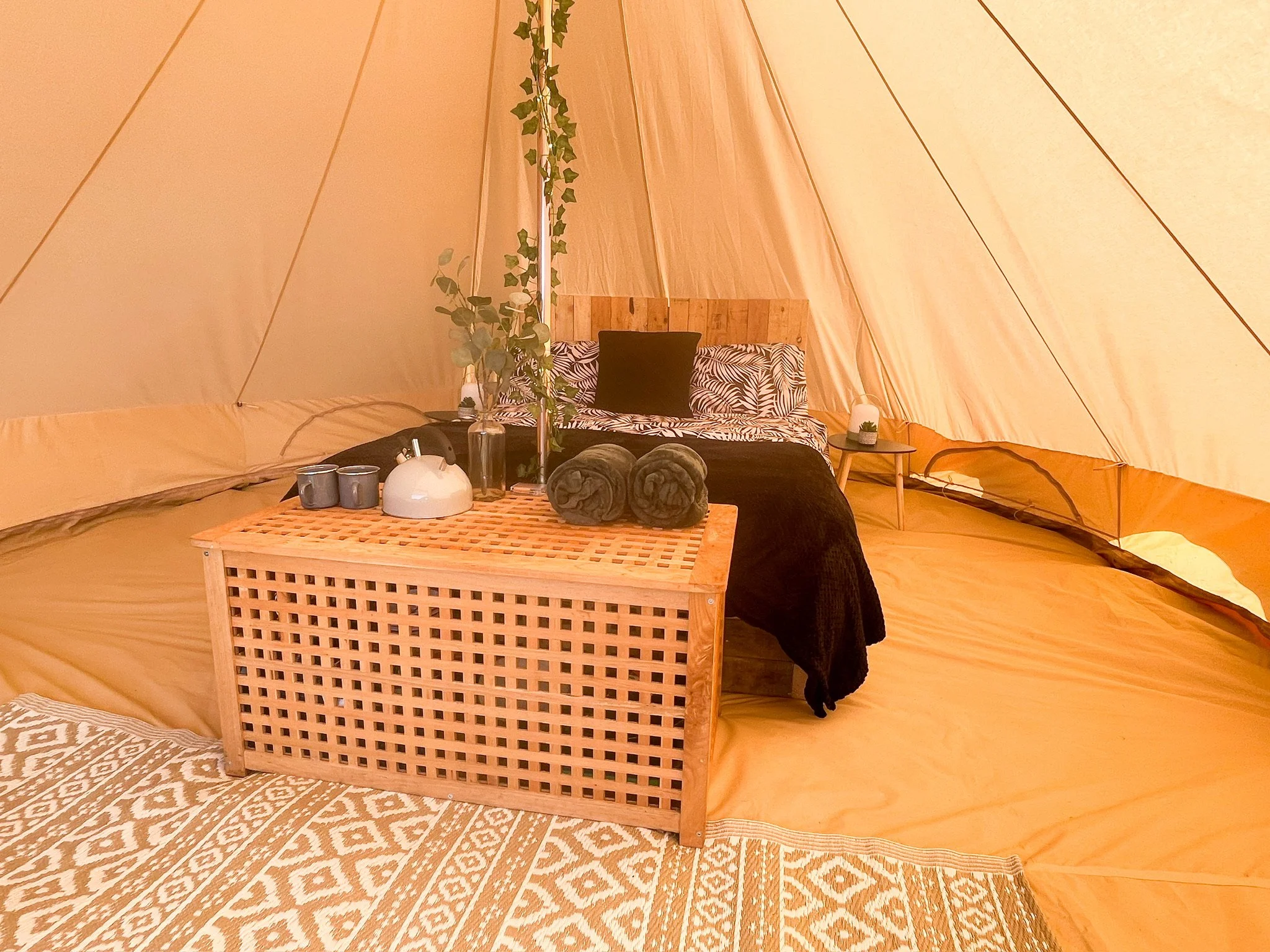 Interior of a cozy camping tent with a bed, two rolled towels, decorative pillows, a side table with a lamp, and a wooden lattice table with cups and a plant.