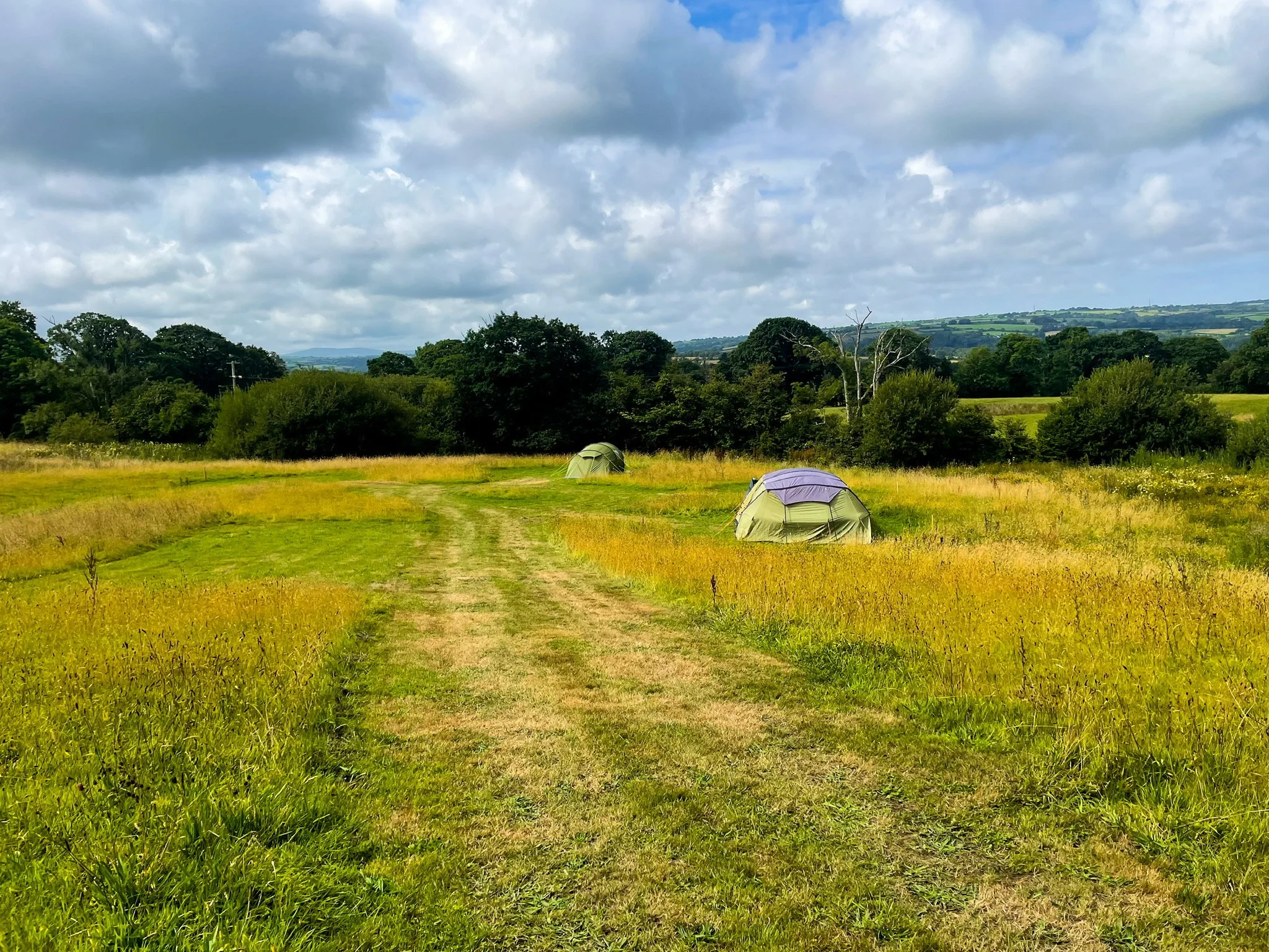 A grassy field with two tents set up, surrounded by trees and hills under partly cloudy skies.