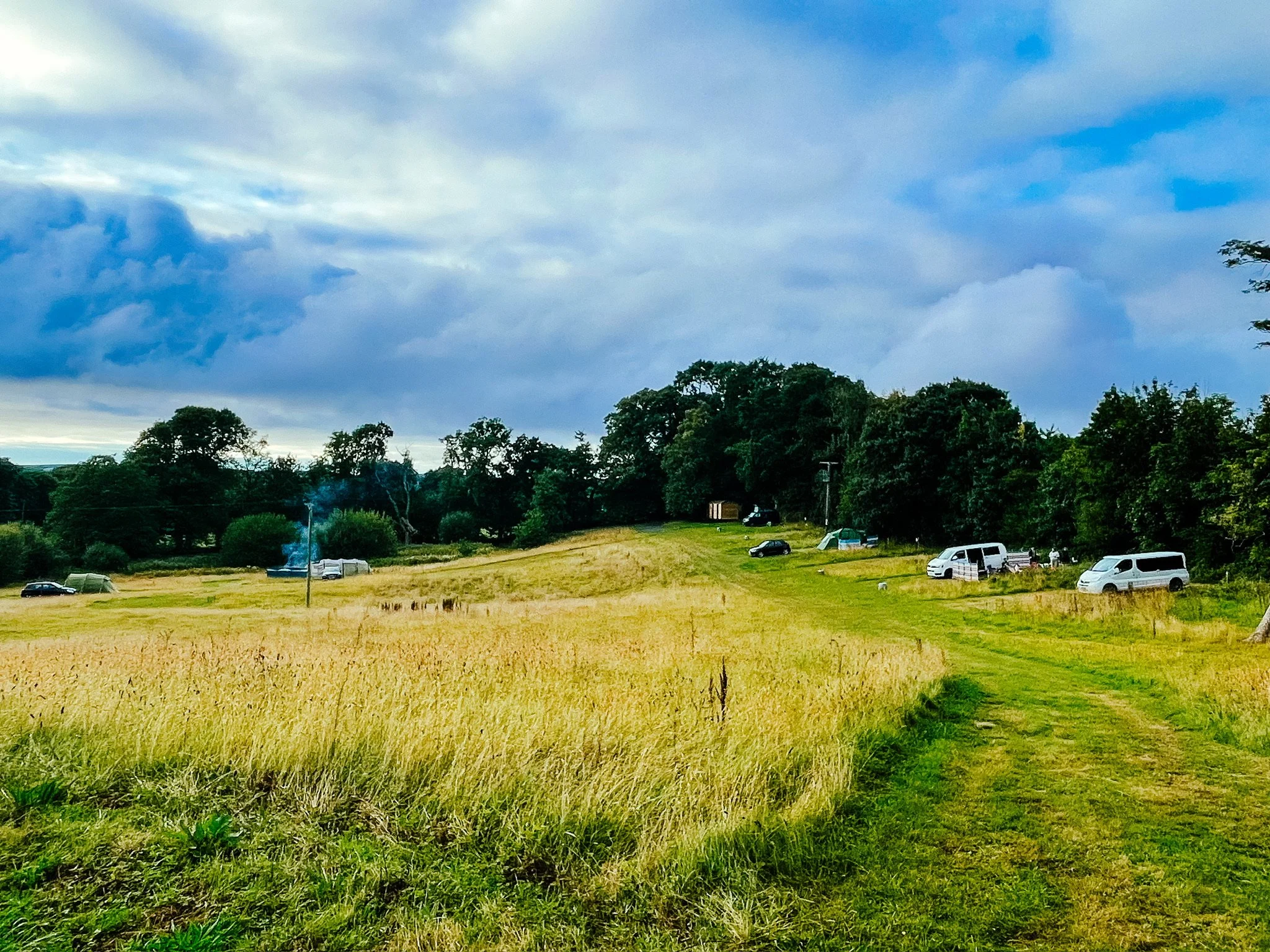A grassy field with a dirt path leading uphill, surrounded by trees and parked vehicles under a partly cloudy sky.