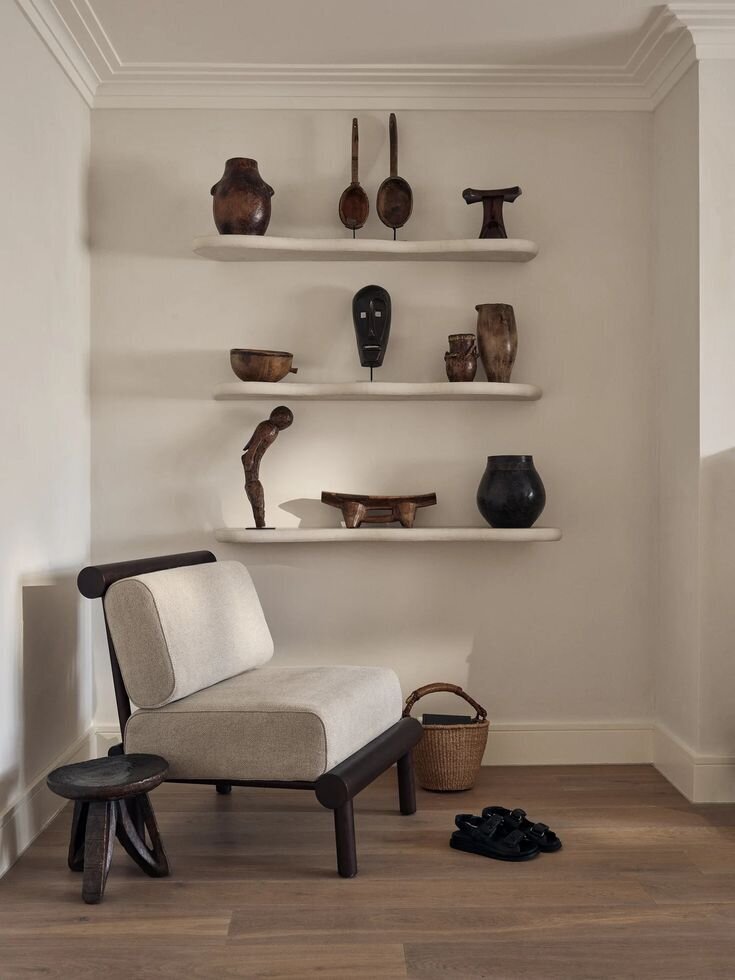 Interior of a room with three white shelves holding wooden and ceramic artifacts, and a beige armchair with black wooden frame near the wall, a small dark wooden stool, a woven basket on the floor, and a pair of black sandals.