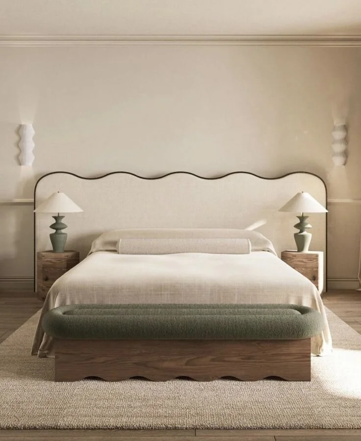 A neatly made bed with a cream bedspread, a rectangular bolster pillow, and a wavy-edged headboard. There are two wooden nightstands with matching lamps on each side of the bed. A green upholstered bench is at the foot of the bed, all placed on a beige textured rug in a minimalist bedroom.