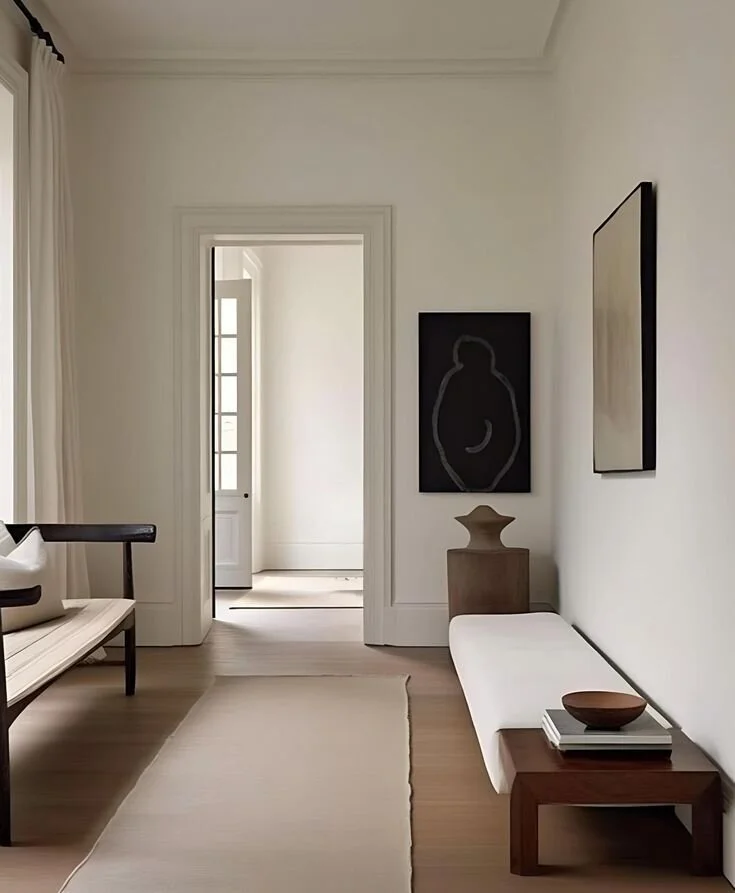 Minimalist living room with white walls, beige rug, wooden bench with pillow, white cushion on a wooden bench, abstract artwork on wall, small wooden side table, and doorway leading to a balcony or another room.