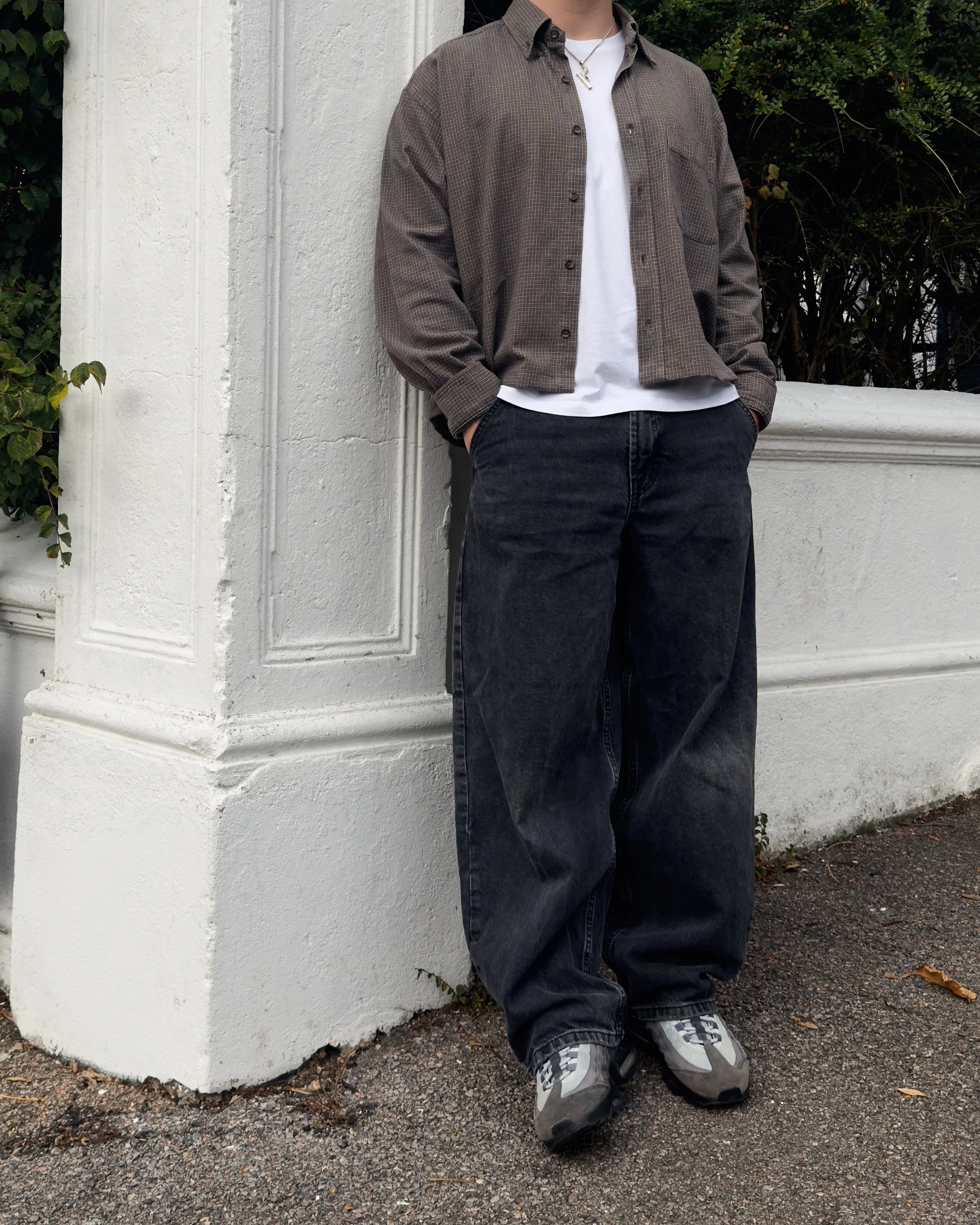 Person wearing a brown shirt, white t-shirt, dark jeans, and sneakers leaning against a white wall.