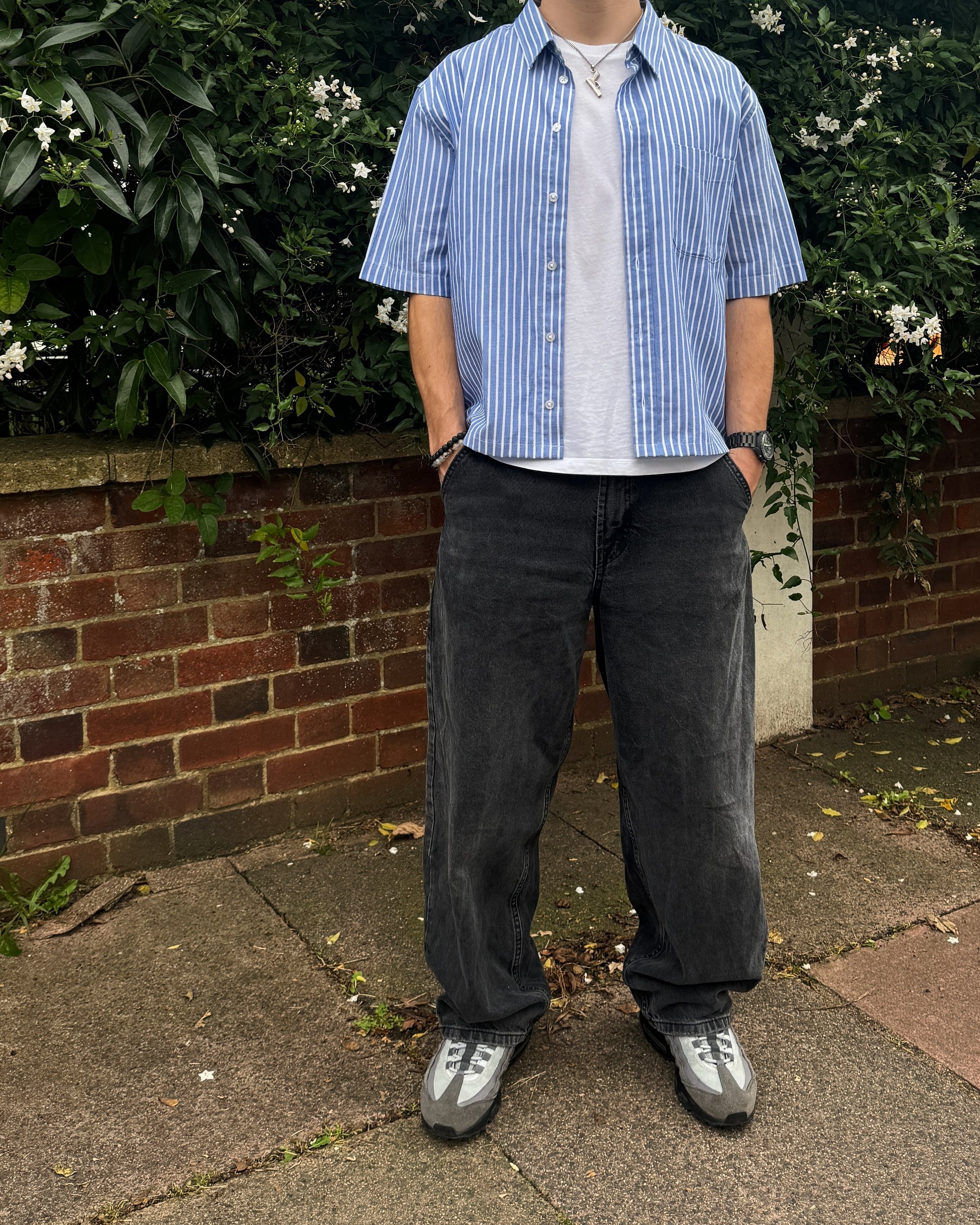 Person wearing a blue striped shirt, white T-shirt, dark jeans, and gray sneakers, standing on a sidewalk with a brick wall and greenery in the background.