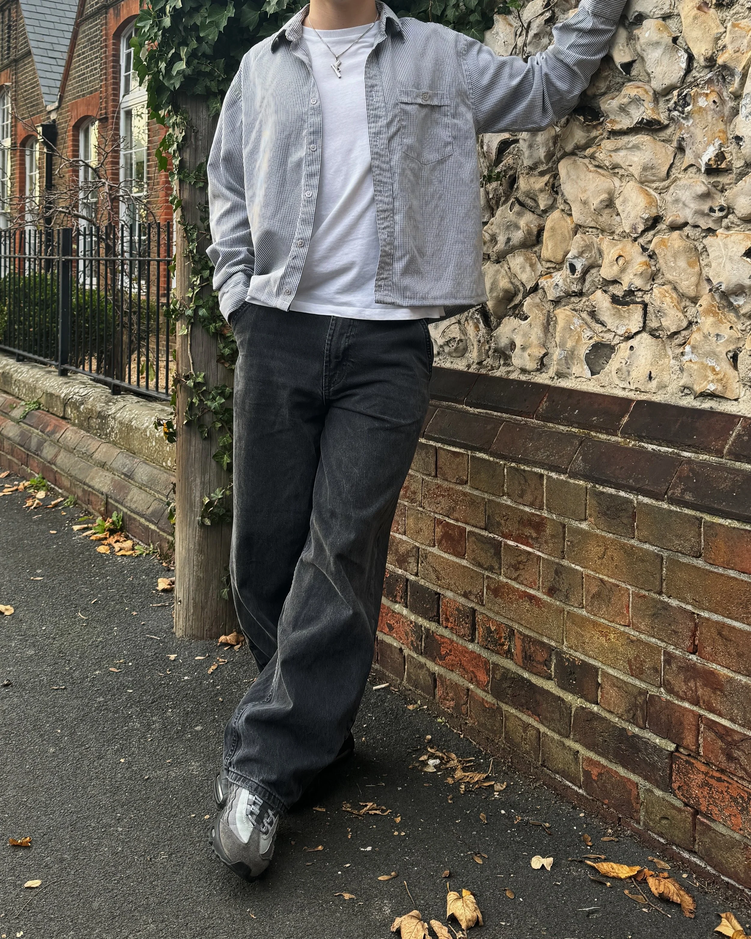 Person leaning against a rock wall, wearing a striped shirt, white T-shirt, dark jeans, and sneakers. Background includes ivy and a brick building.