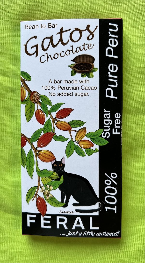 Packaging of a 100% chocolate bar with a drawing of a black cat behind a drawing of a cocoa tree and the words “bean to bar”, “Gatos chocolate”, "a bar made with 100% Peruvian Cacao No added sugar", “feral…just a little untamed” and “100%”.