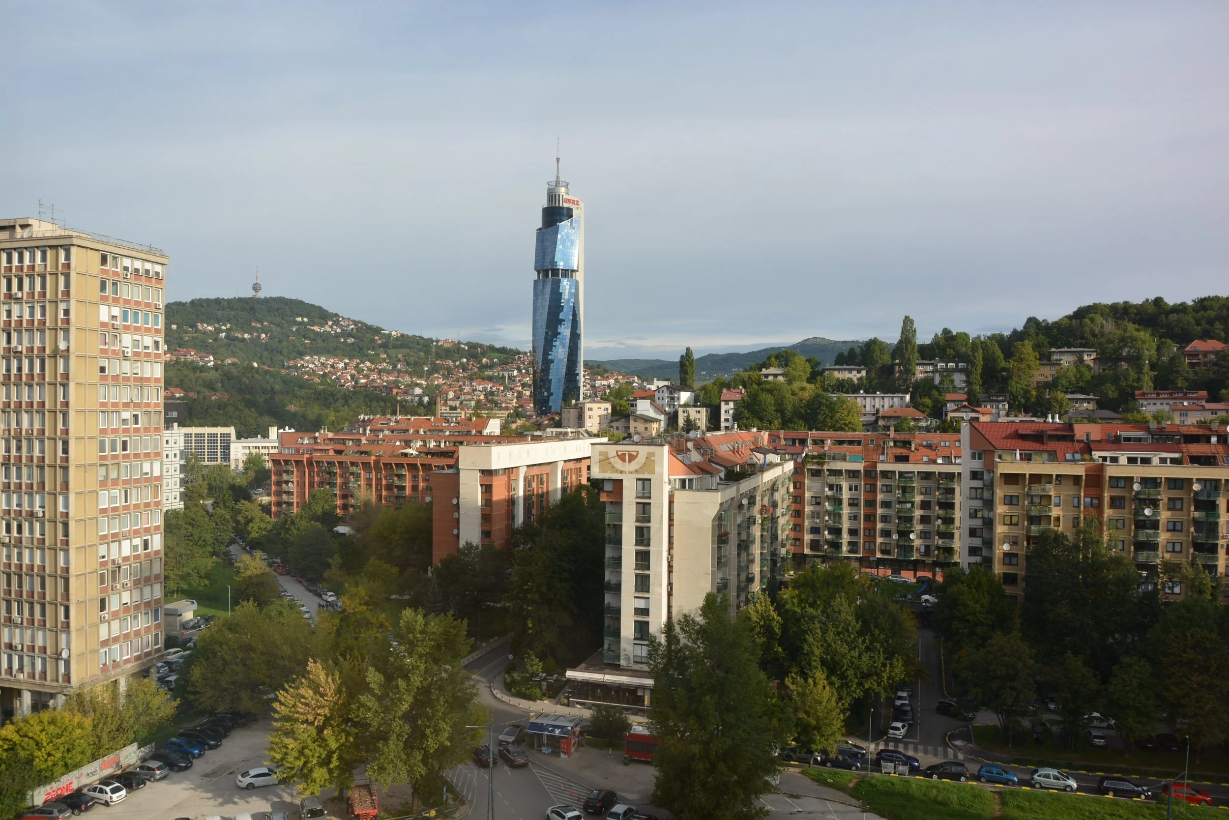 Cityscape with multiple residential buildings, a prominent blue skyscraper, trees lining the streets, and green hills in the background under a partly cloudy sky.