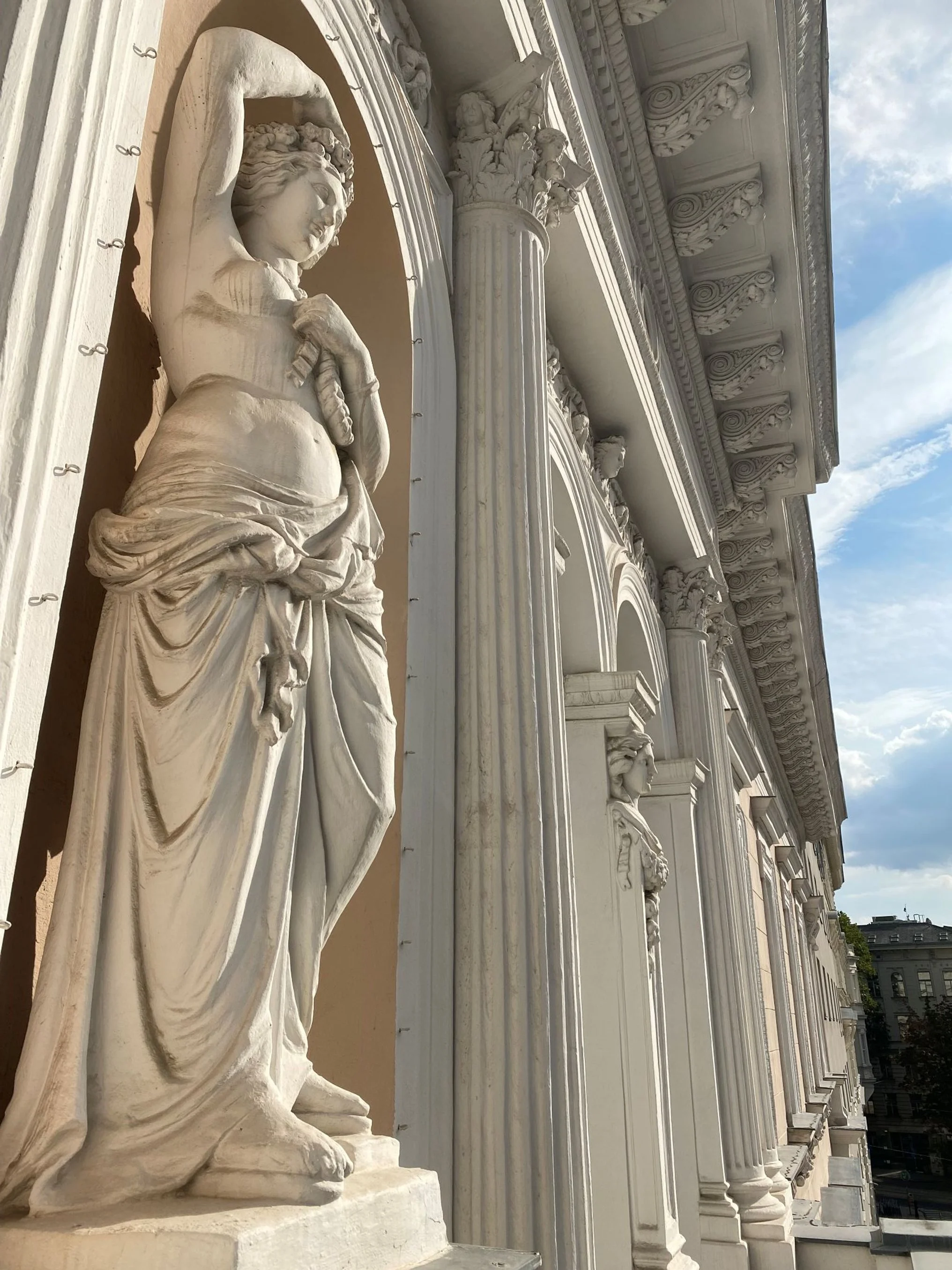 White marble statue of a woman holding a sword, standing against a building with ornate columns and decorative details, under a partly cloudy sky.