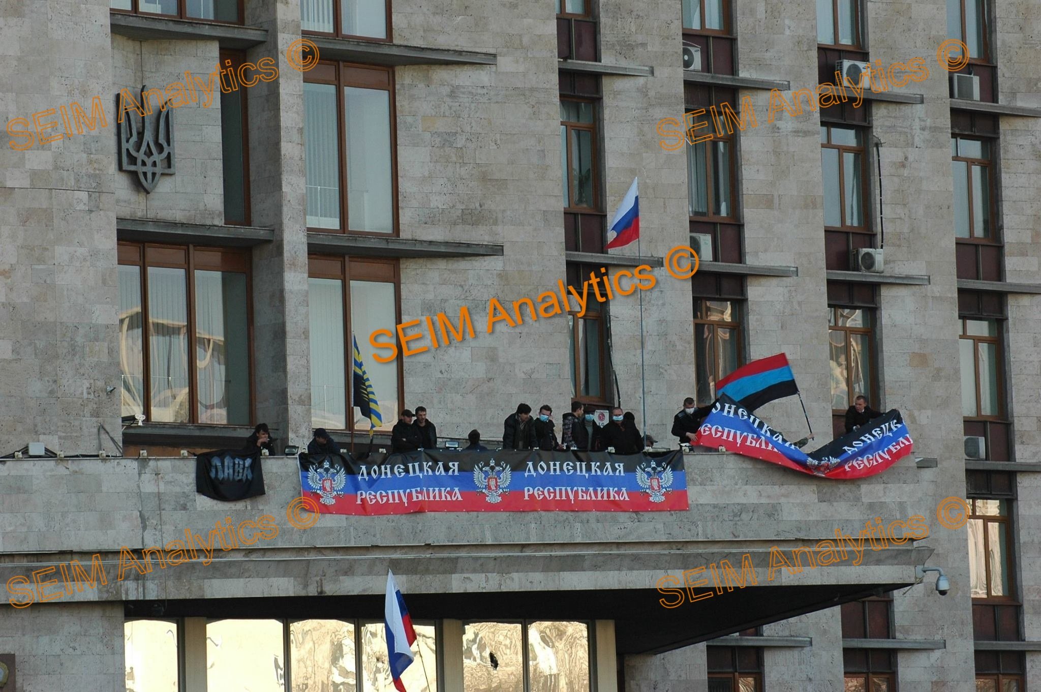 Protest on a building balcony featuring people waving flags, including the Russian flag, and banners with Cyrillic script in Cyrillic and English, with a Ukrainian trident symbol on the building.