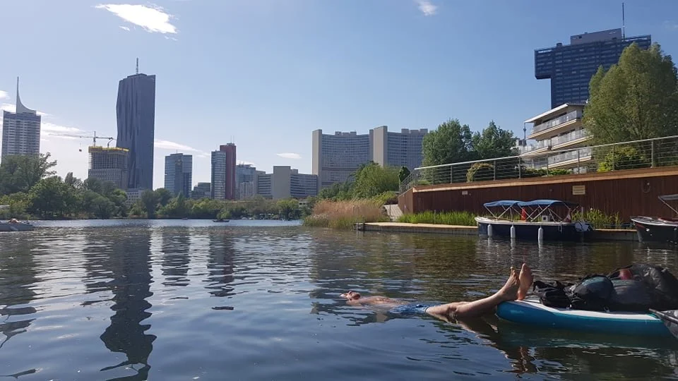 Person floating on a paddleboard in a river with a city skyline in the background, including tall buildings and trees.