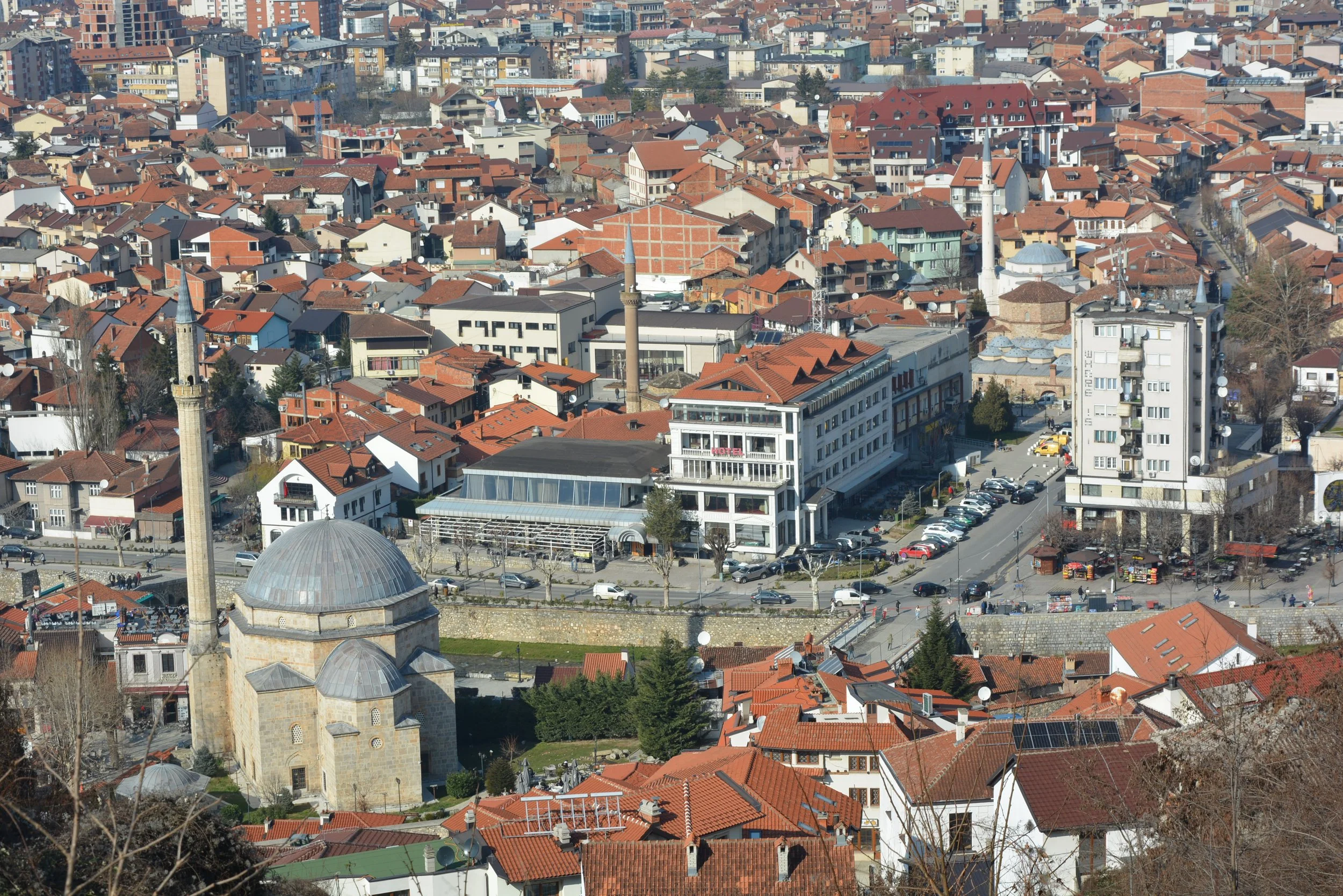 Aerial view of a city with a mix of modern and historic buildings, including a historic mosque with a large dome and minaret, surrounded by residential and commercial structures with red-tiled roofs.
