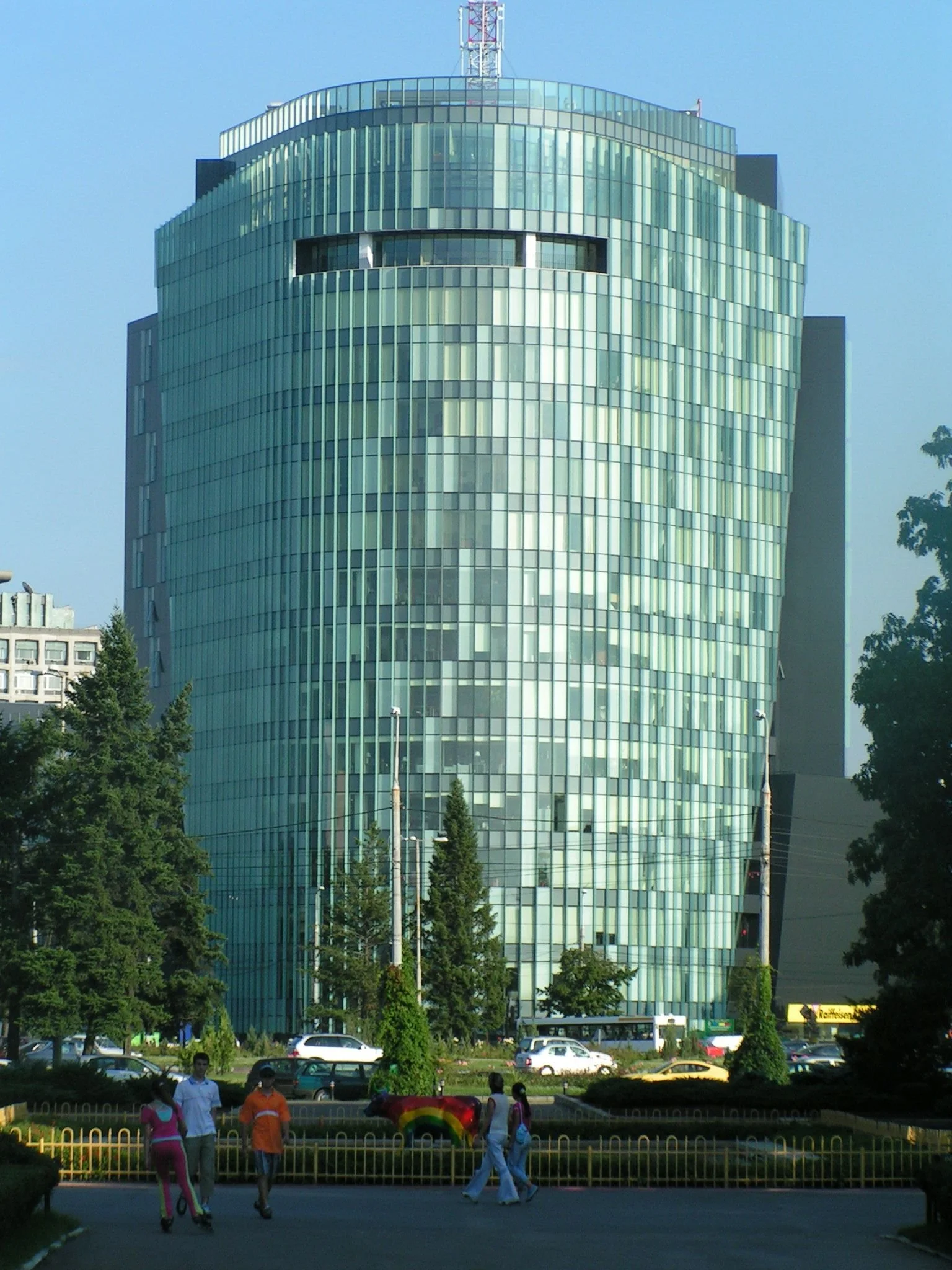 Tall glass skyscraper with a rounded top and modern architectural design, with trees and pedestrians in the foreground.
