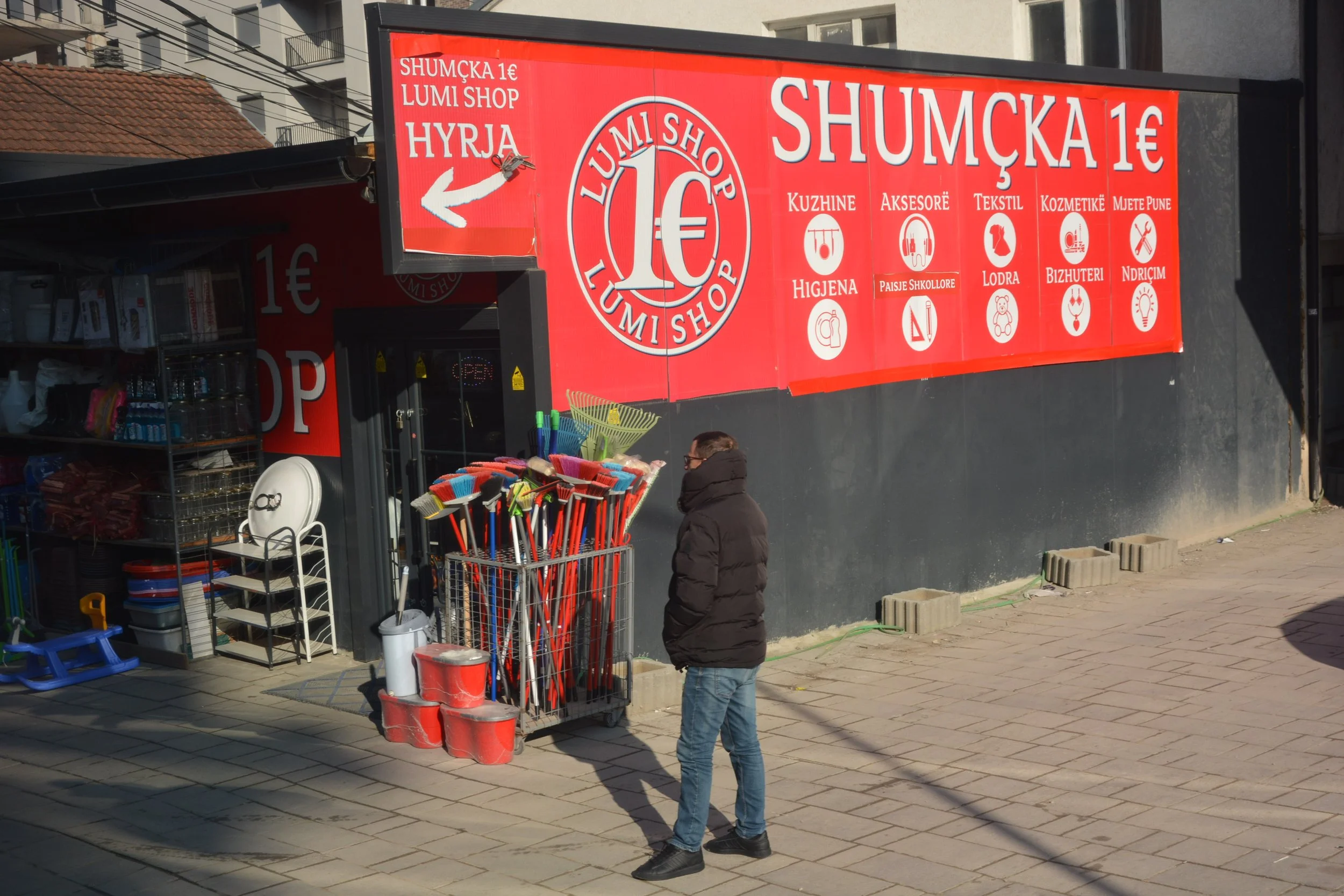 A man standing outside a store with a bright red sign advertising items for 1 Euro. The store sells household and personal care items like gloves, cleaning supplies, and cosmetics. There are various garden tools and cleaning equipment displayed outsi