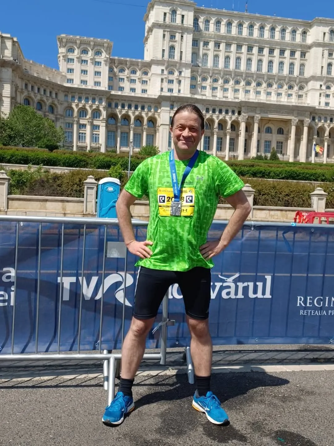 Man standing in front of a large building during a race, wearing a green athletic shirt, black shorts, blue running shoes, and a medal around his neck, with a blue fence and blue portable toilet behind him and a blue sky above.