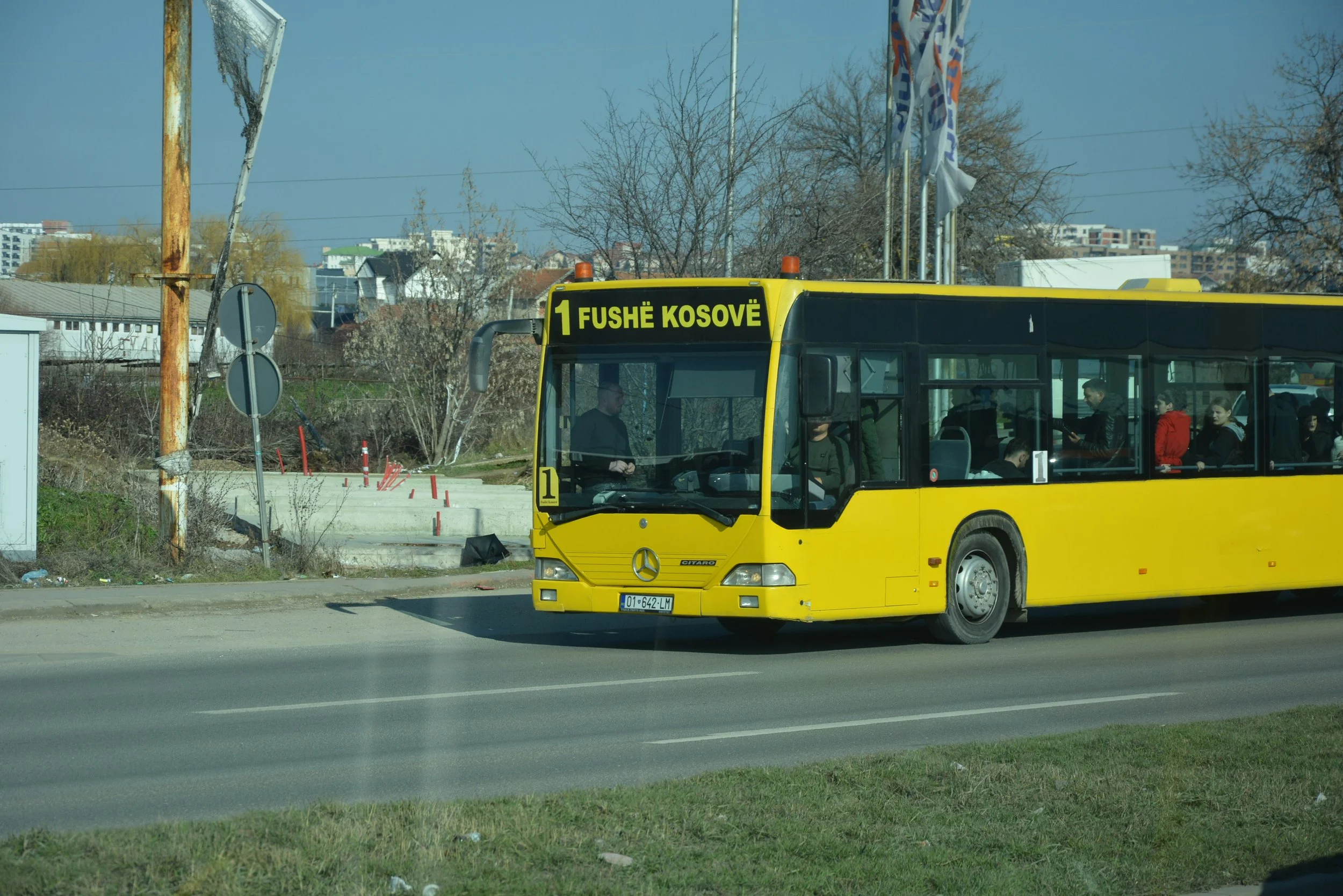 A yellow bus on the road with a digital sign displaying 'Fushë Kosovë'. There are passengers visible inside the bus, and the background shows trees, buildings, and a cloudy sky.