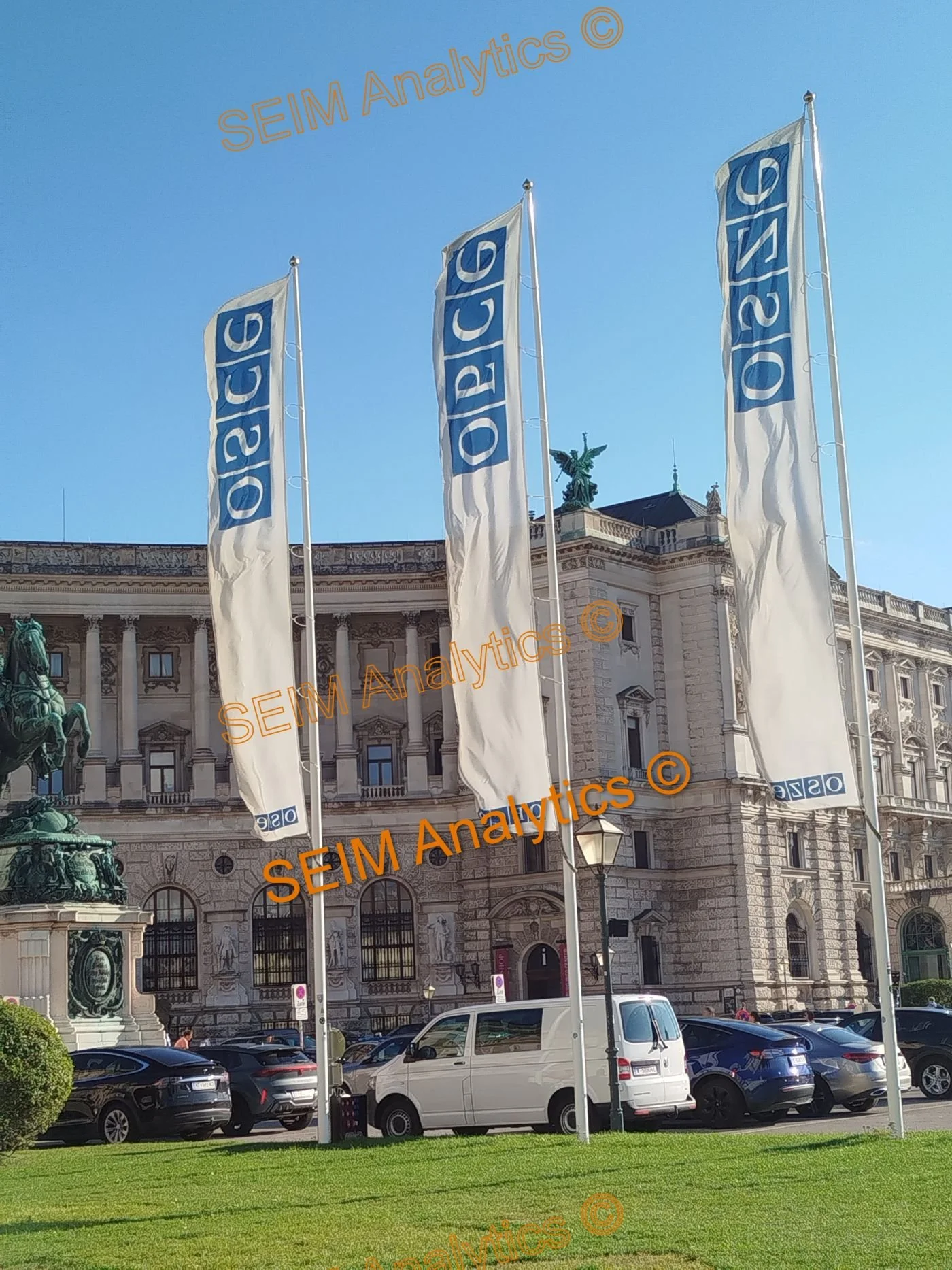 Three large flags with the OSEC logo on a historic building with statues and a parked white van in front. The sky is clear and blue.