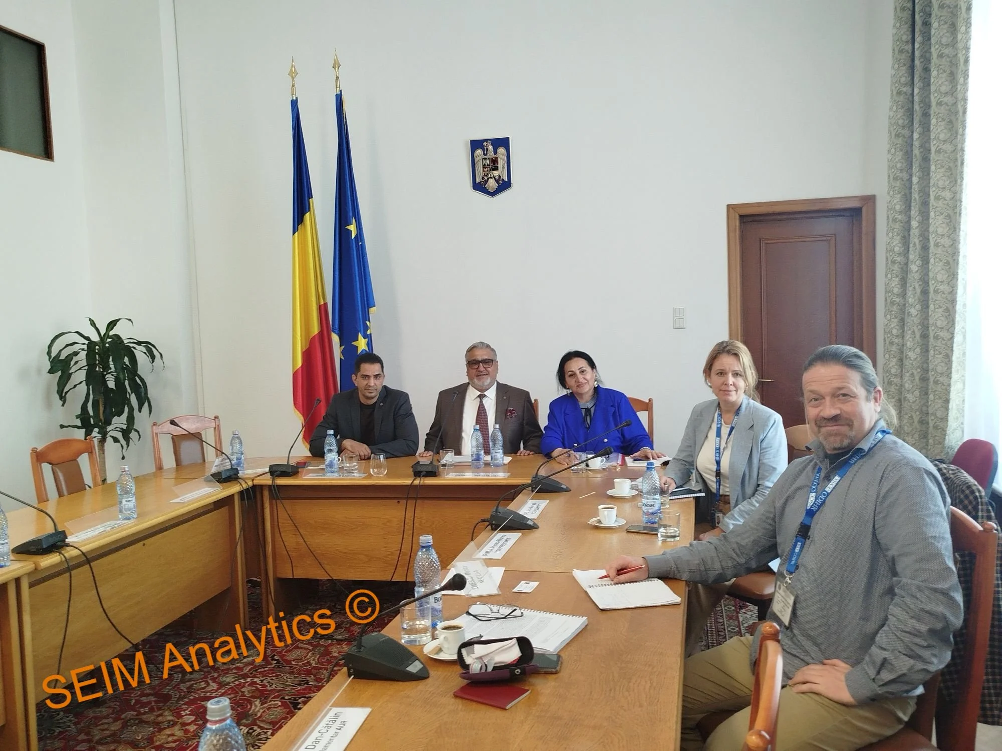 Group of five people sitting around a conference table in a meeting room with flags behind them.