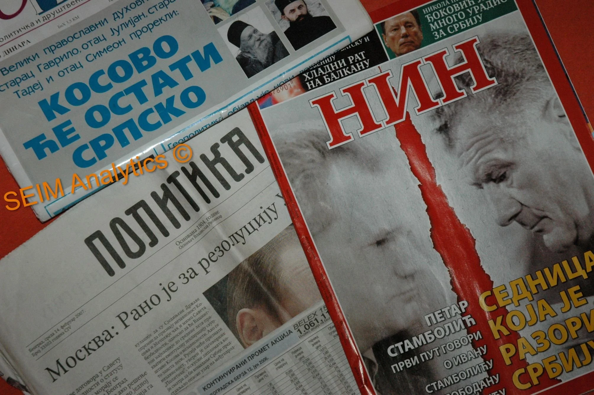 Several political newspapers in Serbian, with one showing a close-up of Vladimir Putin's face and a red vertical stripe, another with a headline in blue text, and a third with a black headline and some smaller images of people.