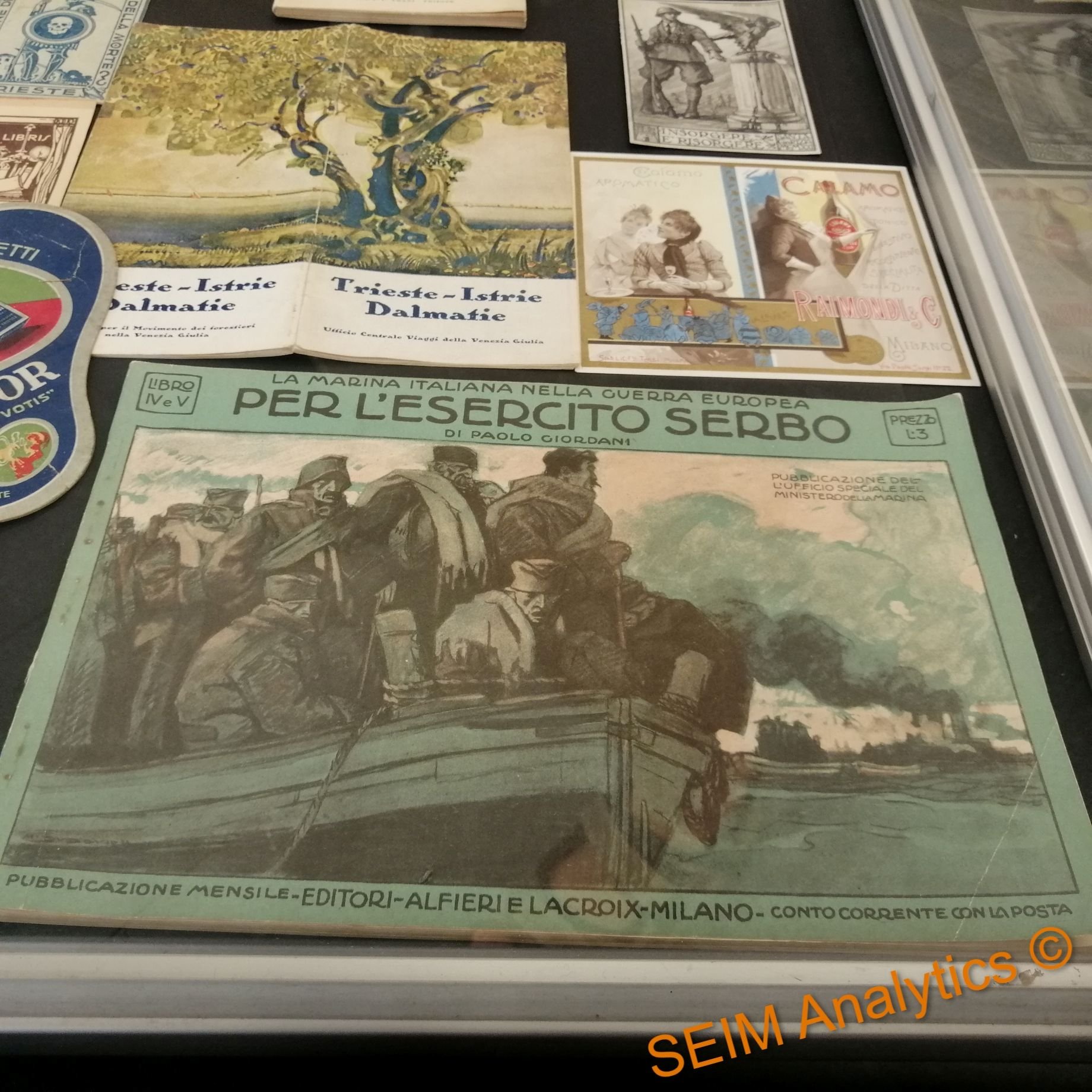 A display case with vintage posters and flyers, including a green Italian military publication titled 'Per l'Esercito Serbo' featuring soldiers on a boat during World War I.