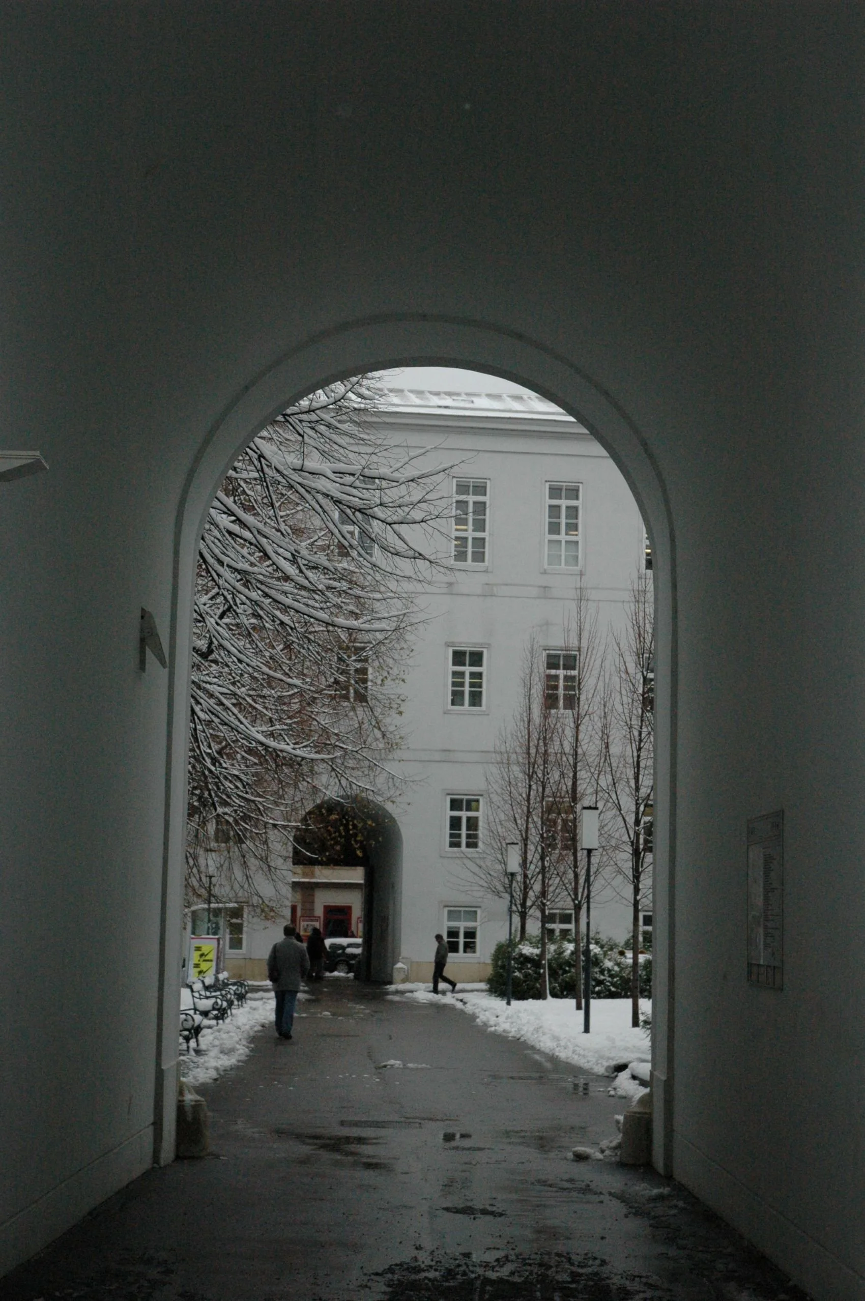 View through a white archway of a snowy courtyard with a large white building, leafless trees, and a few walking people.