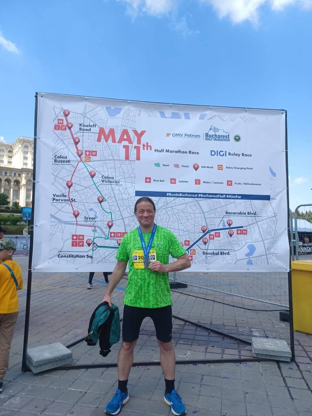 A man in a green running shirt and black shorts stands in front of a large race map poster, holding a medal, at a marathon event in Bucharest, Romania. The poster displays the race route and various stations along the half marathon and relay race courses.