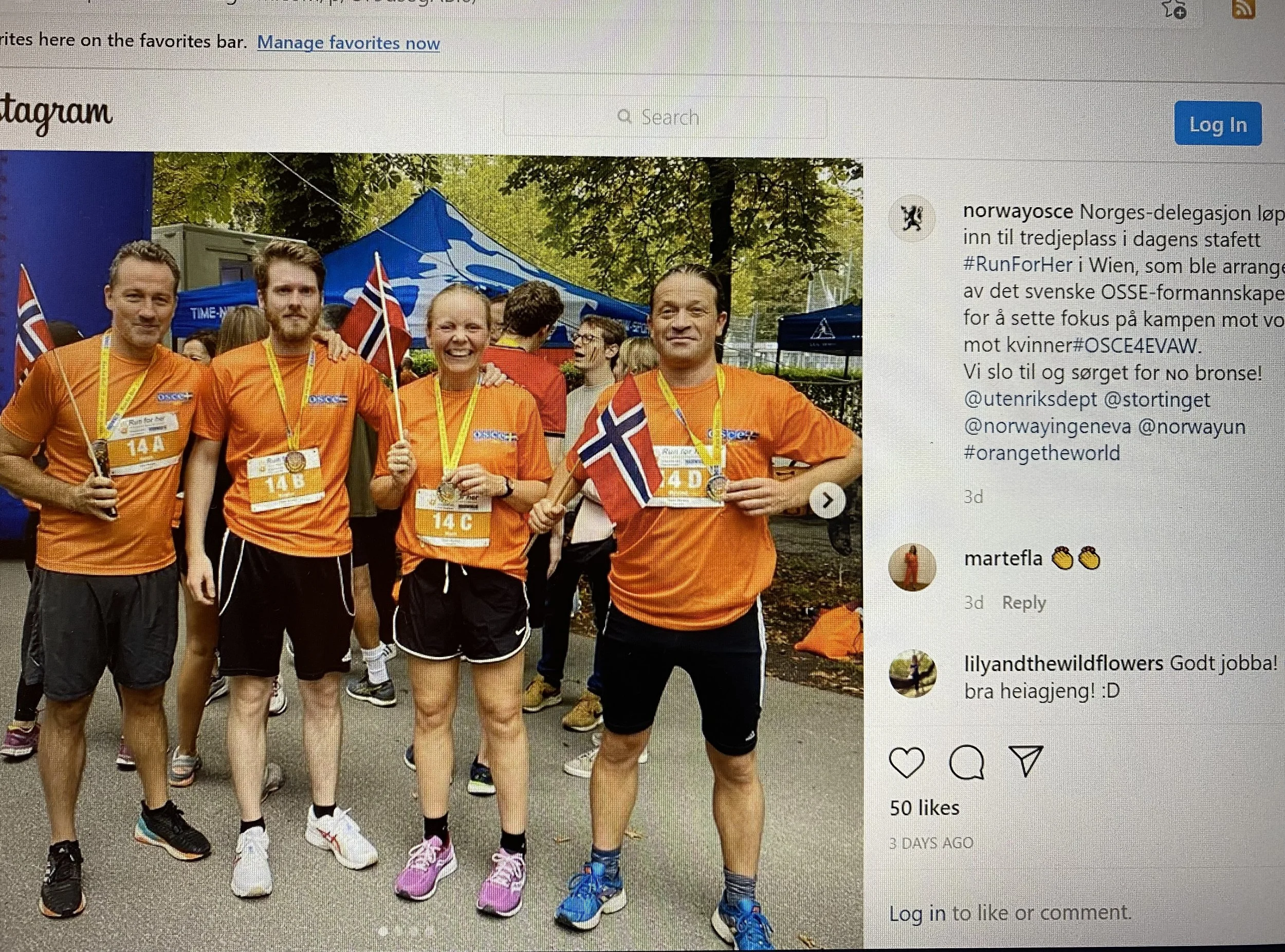 Four people wearing orange running shirts and medals around their necks posing for a photo after a race, some holding small Norwegian flags. They are standing outdoors near trees, with race tents and other participants in the background.