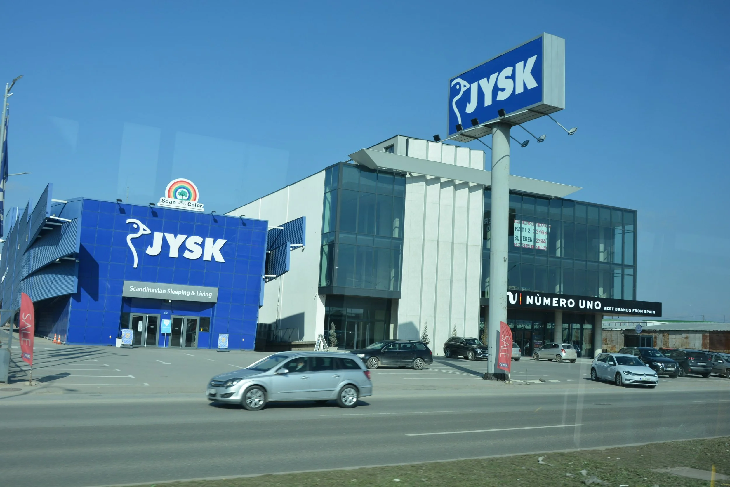 A modern shopping center with a large blue JYSK sign, a JYSK store called 'Scandinavian Sleeping & Living,' a sign for 'NÚMERO UNO,' and parked cars in front under a clear blue sky.