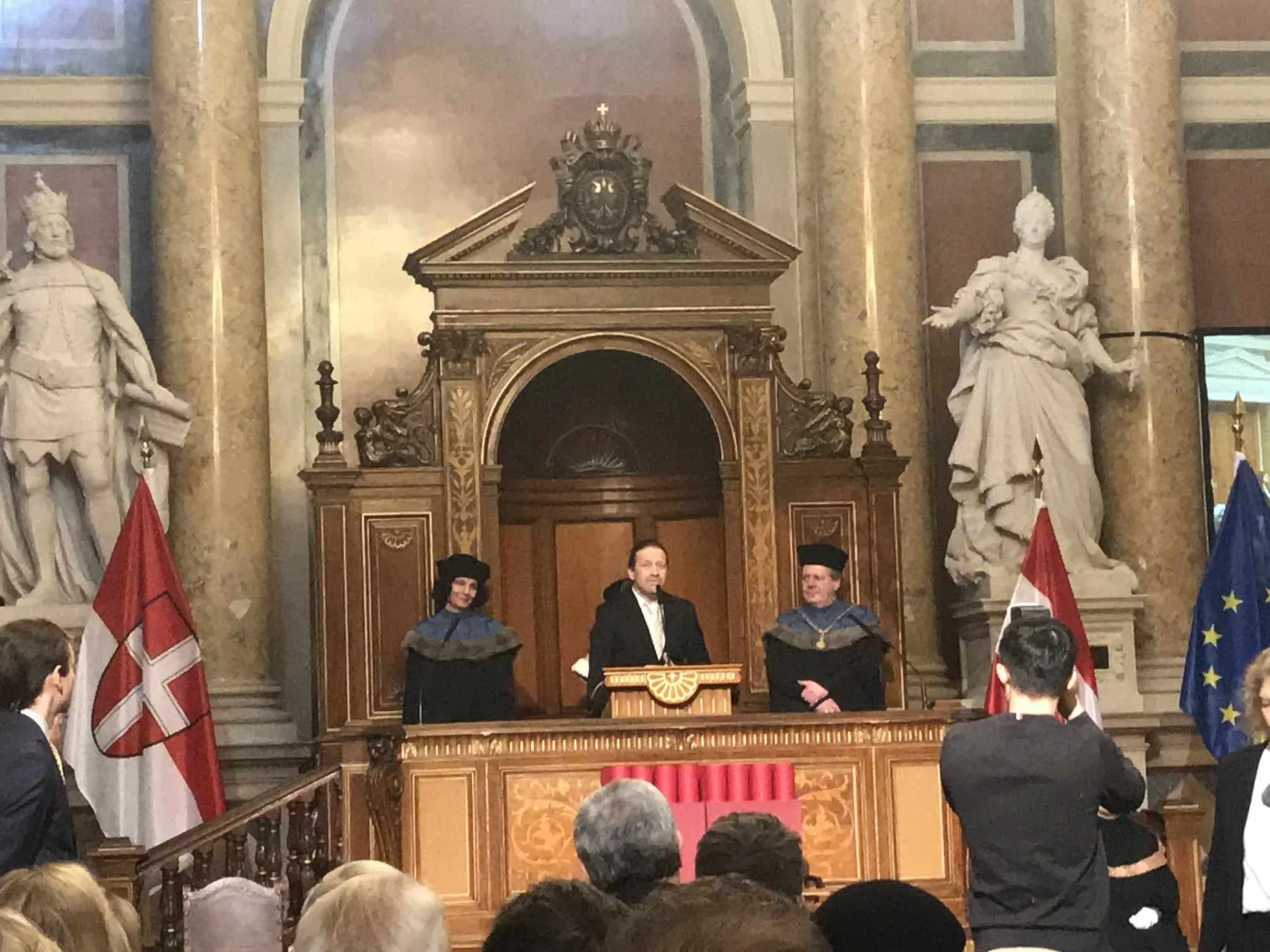 A man standing at a wooden podium in a grand, ornate hall with gold-tiled columns and statues. Two people in academic regalia stand on either side of him. Several audience members are visible, and flags are displayed, including the flags of Denmark, the European Union, and another unidentified flag.