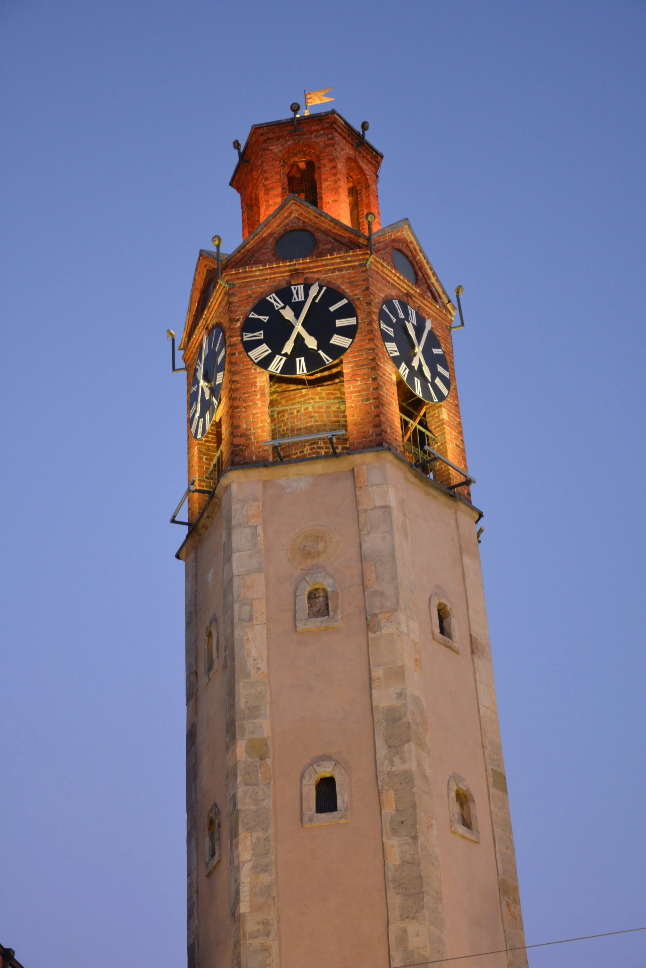 A tall clock tower made of brick and stone with two clock faces displaying approximately 12:03, set against a clear blue evening sky.