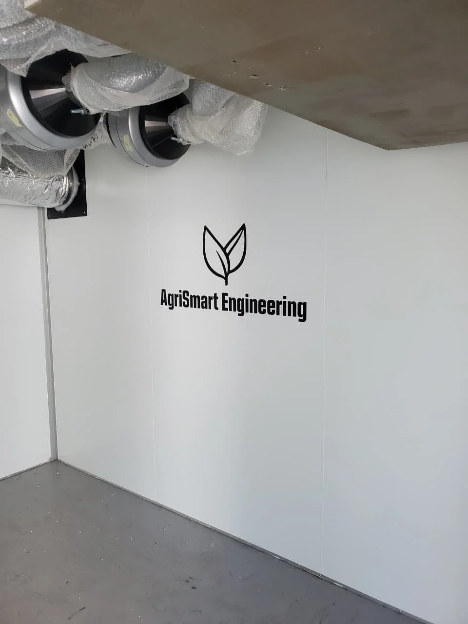 Interior view of a room or construction site with machinery and insulation wrapped pipes, and a wall featuring a logo of a leaf and the text 'AgriSmart Engineering'.