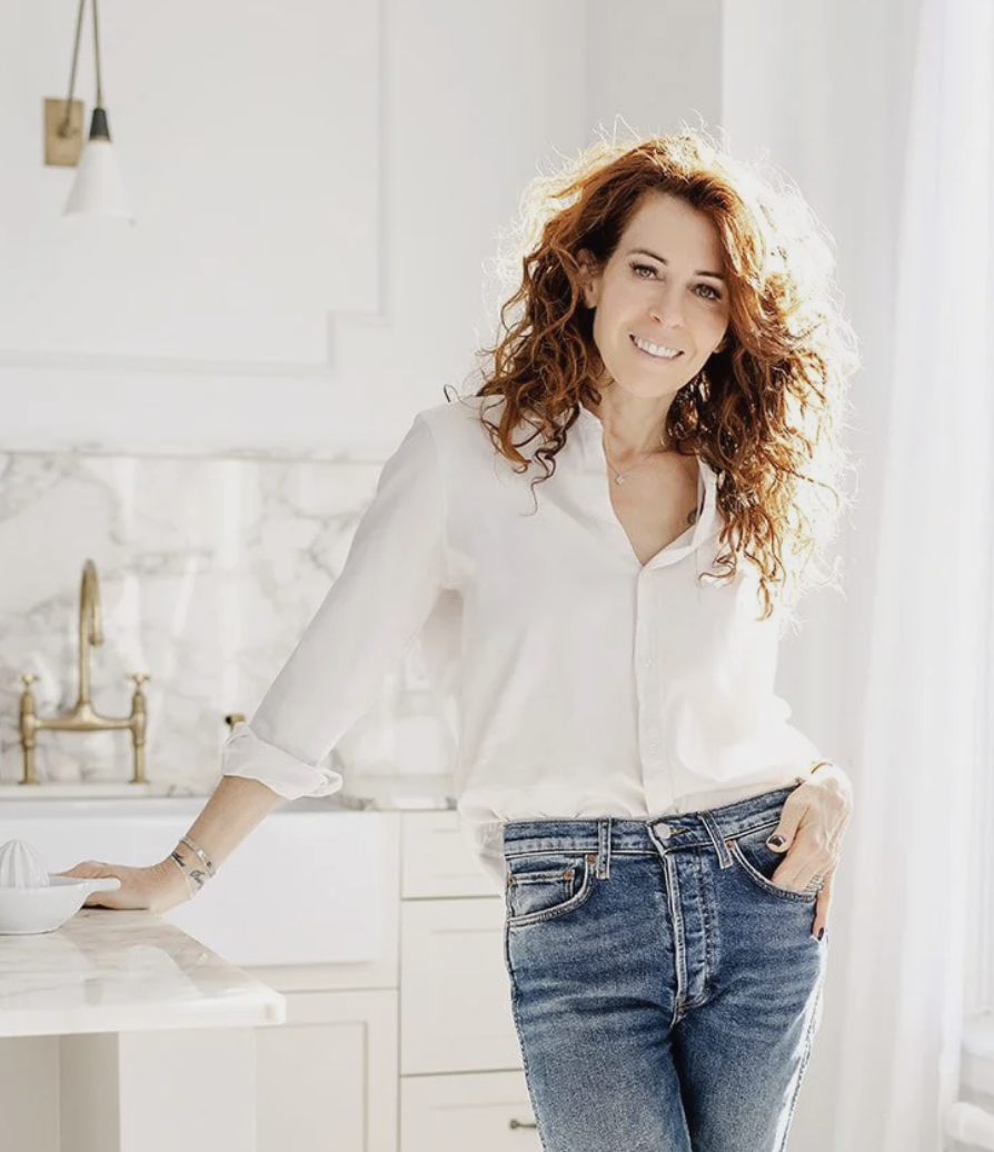 Beautiful slim woman with long curly red hair, standing in a white, modern kitchen wearing jeans, and a white shirt.