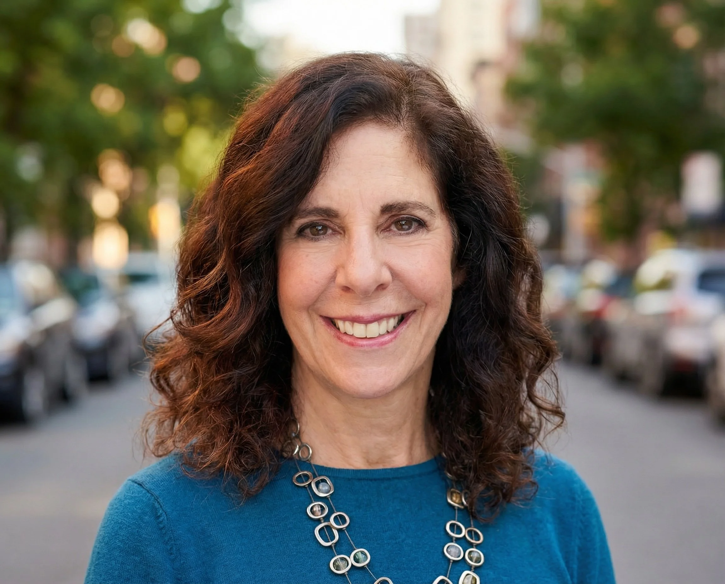 Close-up of a middle-aged woman with brown, curly hair smiling outdoors, wearing a bright blue shirt and silver jewelry, with blurred greenery and parked cars in the background.