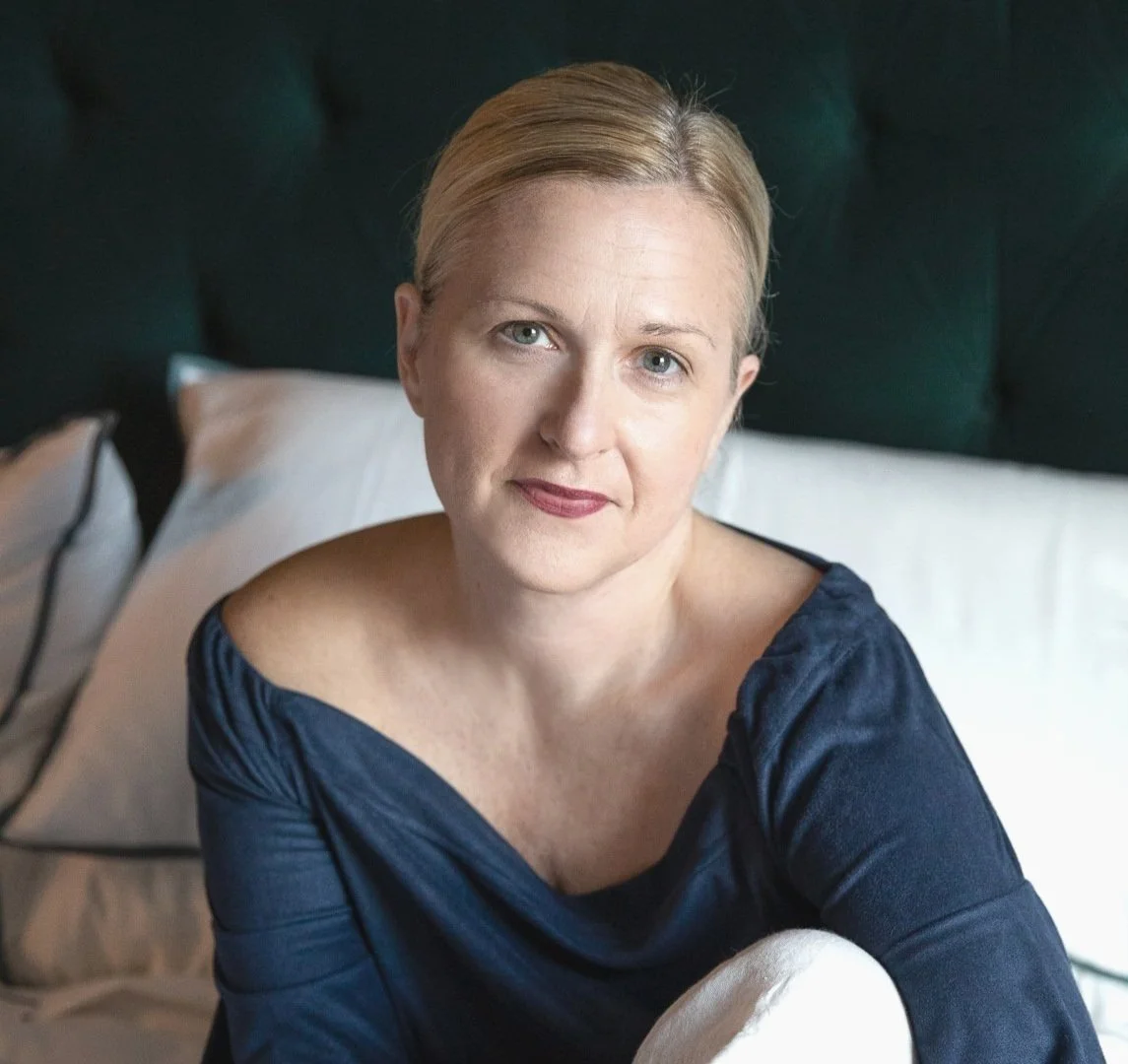 A woman with short blonde hair, wearing a dark blue top, sitting on a bed with white pillows and a dark headboard in the background.
