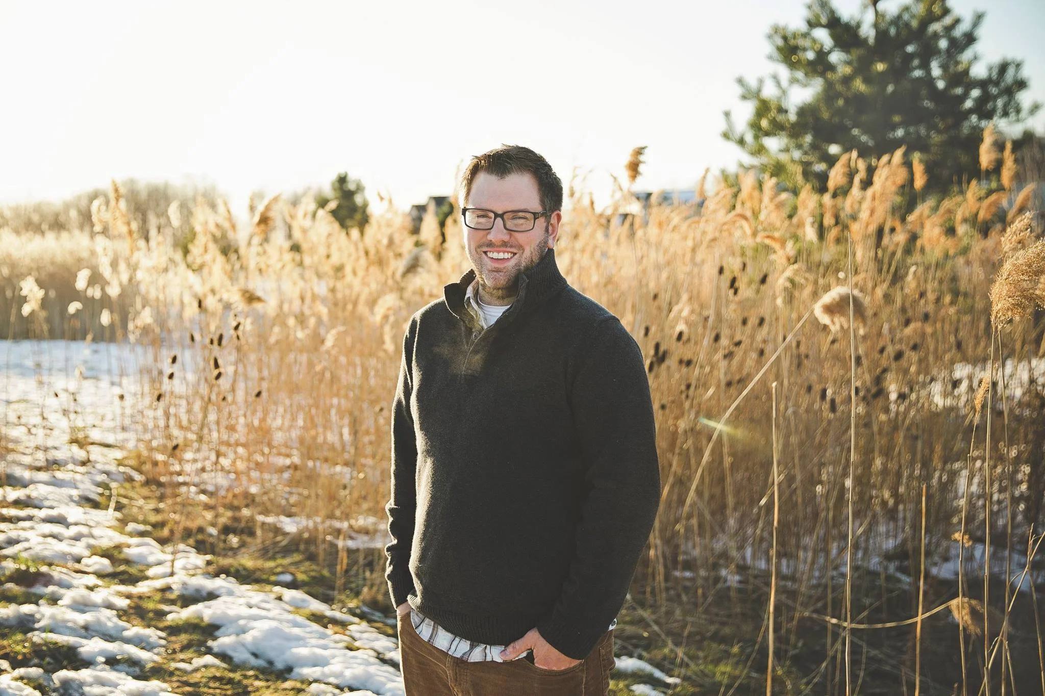 Smiling man with glasses wearing a black jacket standing outdoors in a field with tall grass and snow, sunshine in the background.
