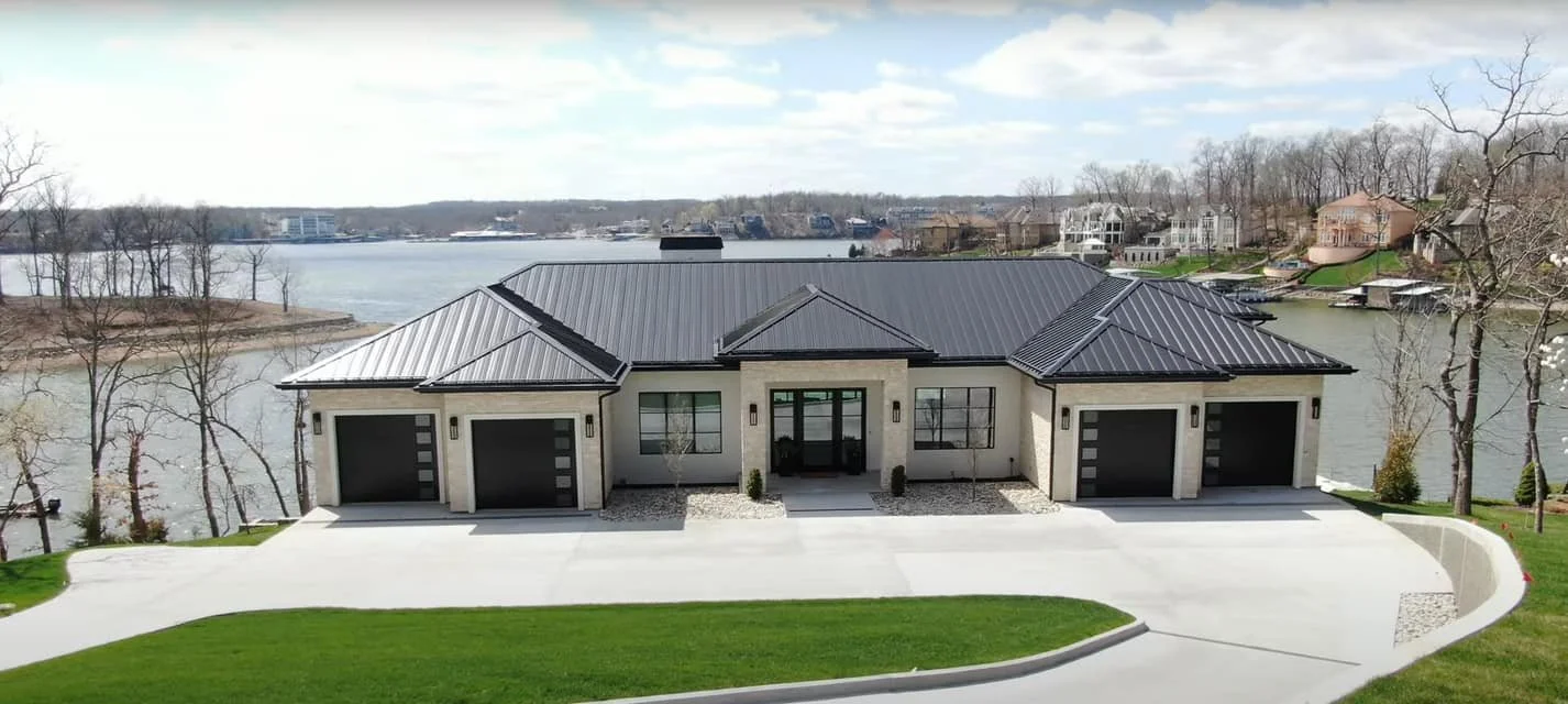 Modern waterfront house with three garages and black metal roof, green lawn, trees, and a lake in the background.