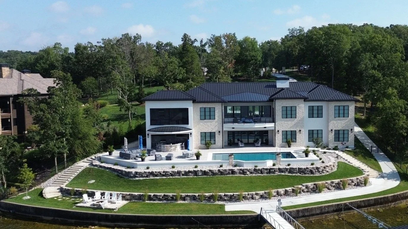 Large white house with black roof, multiple levels, and patio area with swimming pool, outdoor furniture, and landscaped yard, surrounded by trees.